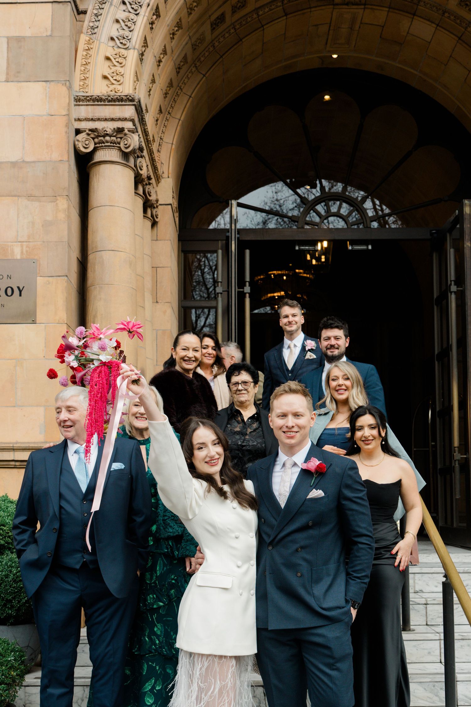 Bride and groom celebrate outside The Savoy hotel with wedding party, bride raising pink bouquet.
