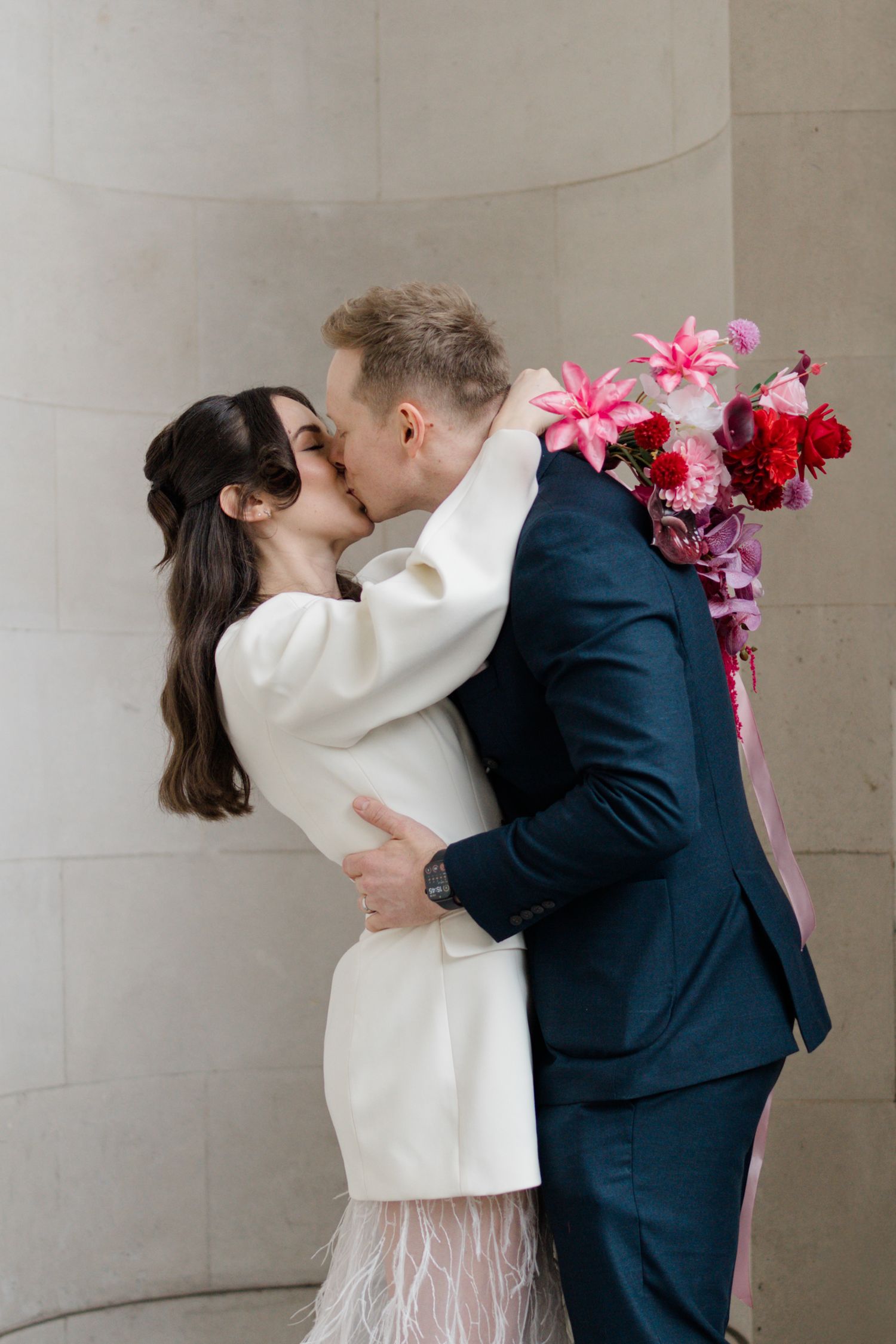 Bride and groom share first kiss outside registry office, bride holding vibrant pink bouquet.
