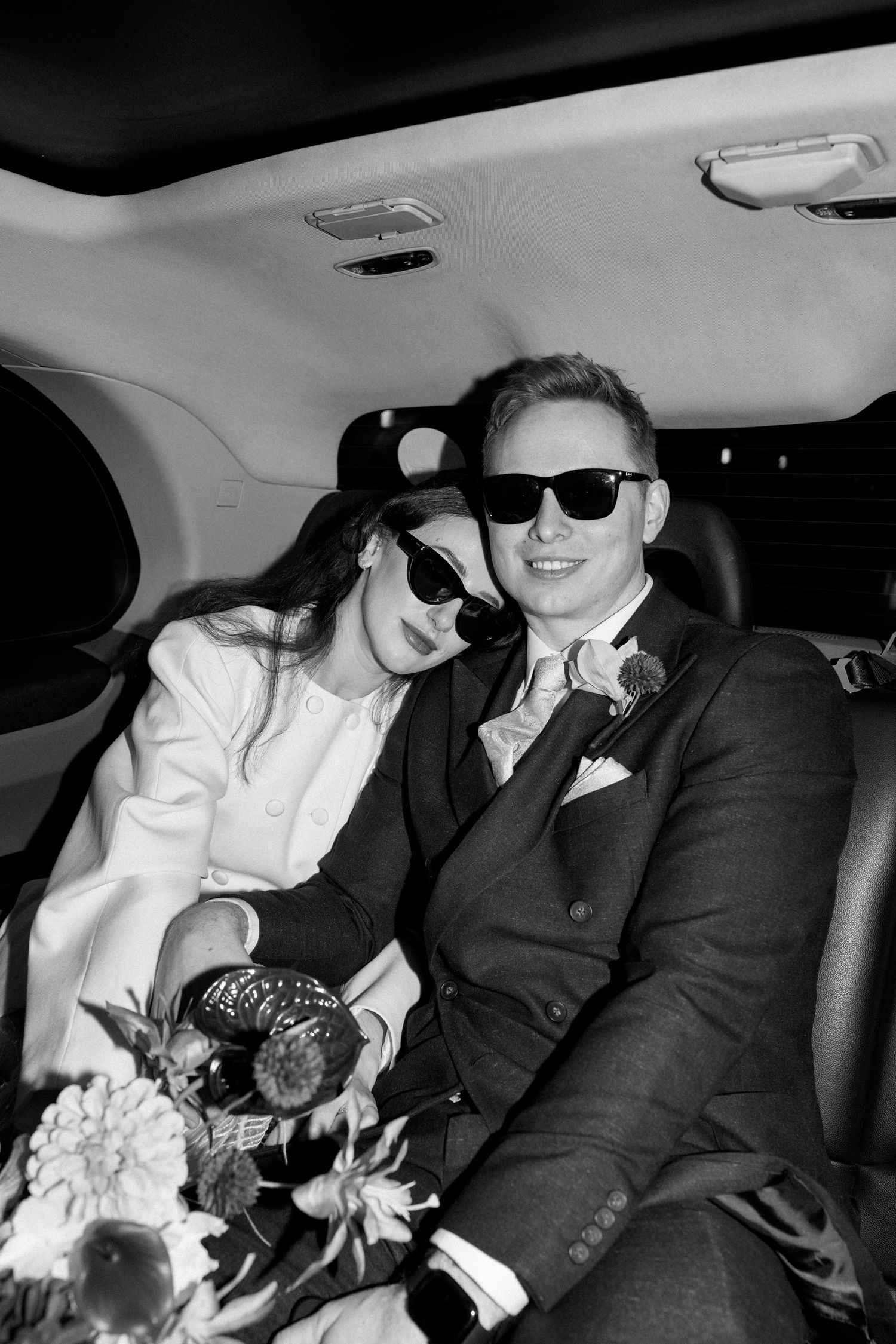 Black and white photo of newlyweds wearing sunglasses, smiling together in a luxury wedding car with bouquet.