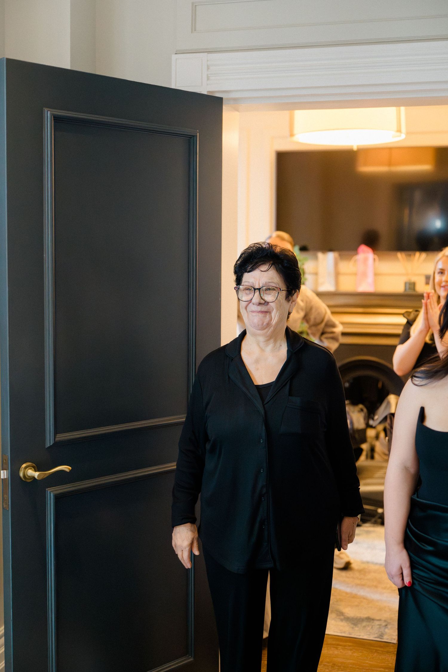 Elderly woman in black outfit smiling while entering a warmly lit room during a wedding celebration.
