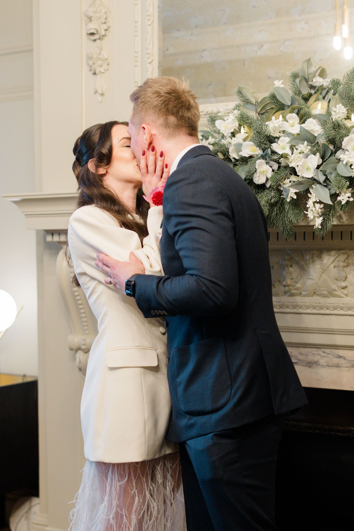 Bride and groom sharing a romantic kiss in front of a floral fireplace mantel at their wedding.