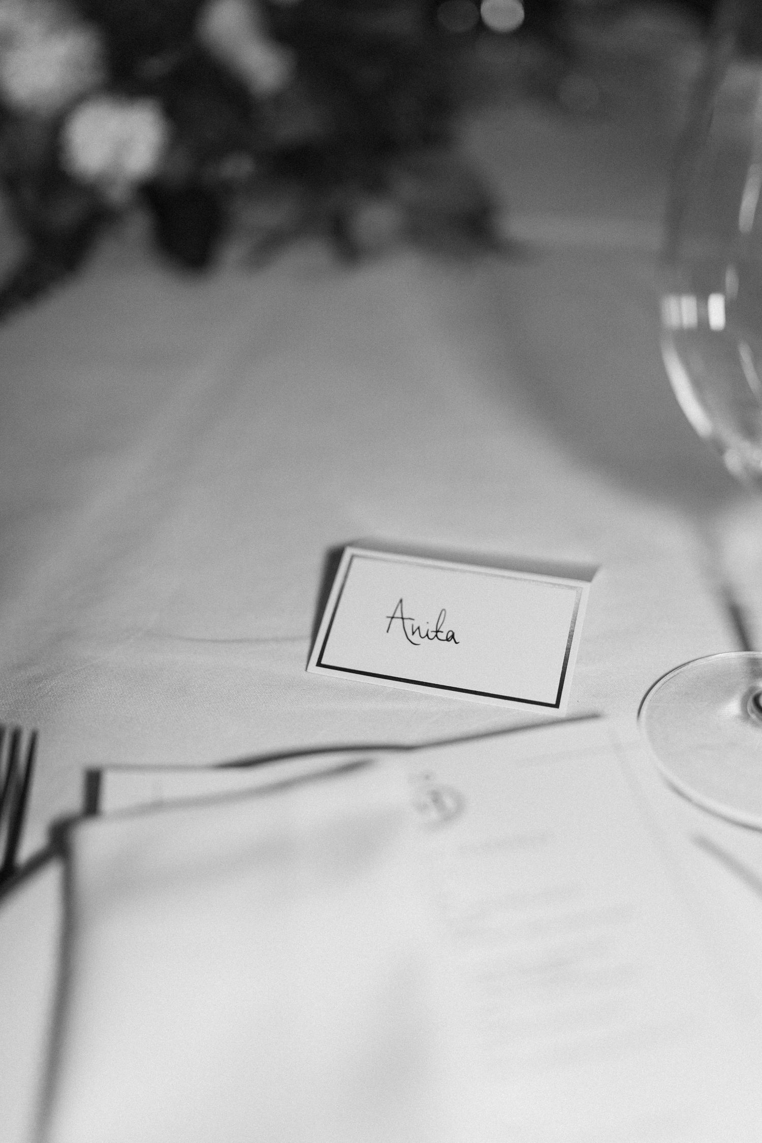 Black and white close-up of an elegant place card reading Anita on a neatly set wedding reception table.