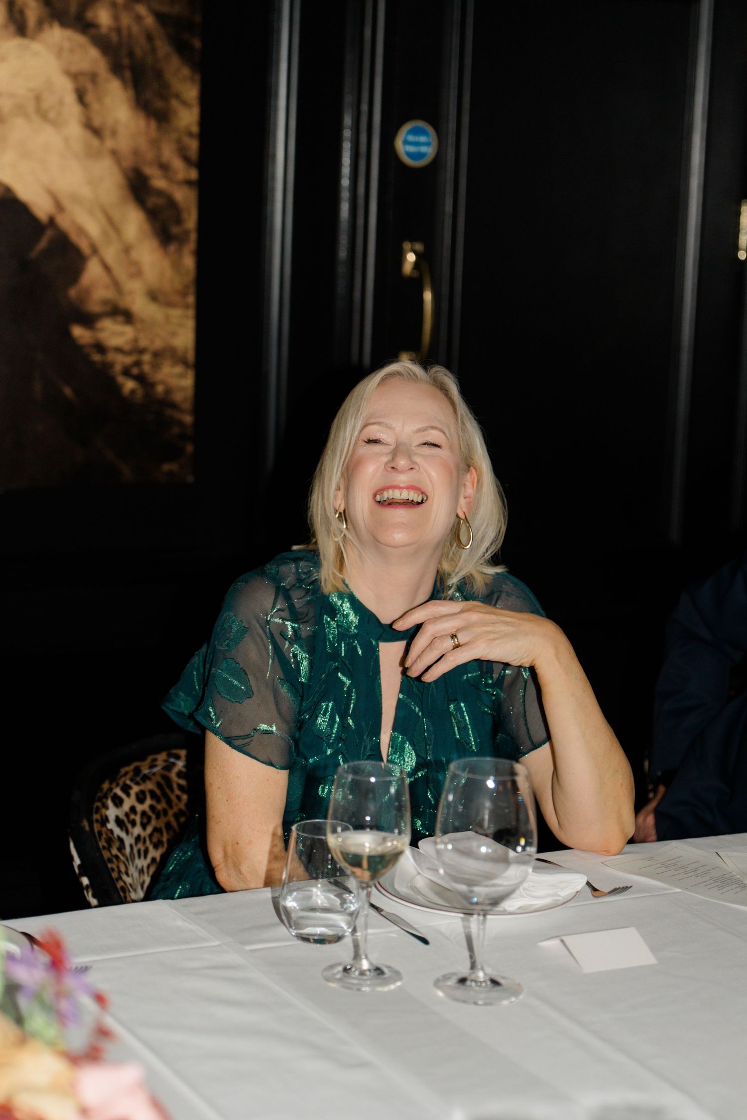 Joyful blonde woman in emerald green dress laughing at wedding reception dinner table with wine glasses.