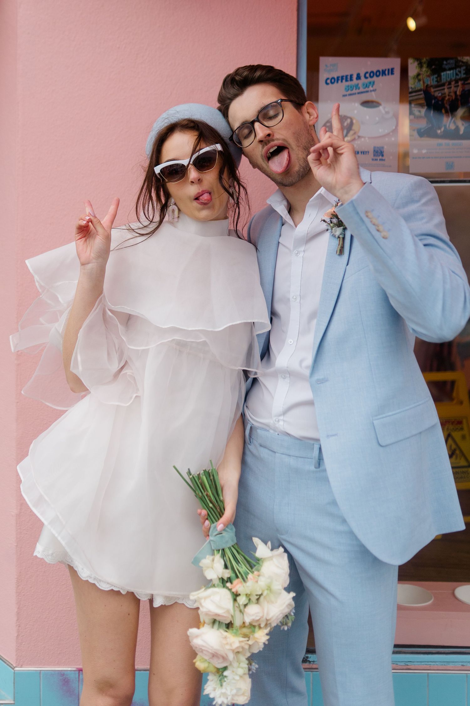 Playful couple in wedding attire, bride in white mini dress with bouquet, groom in light blue suit, posing outside pink storefront.