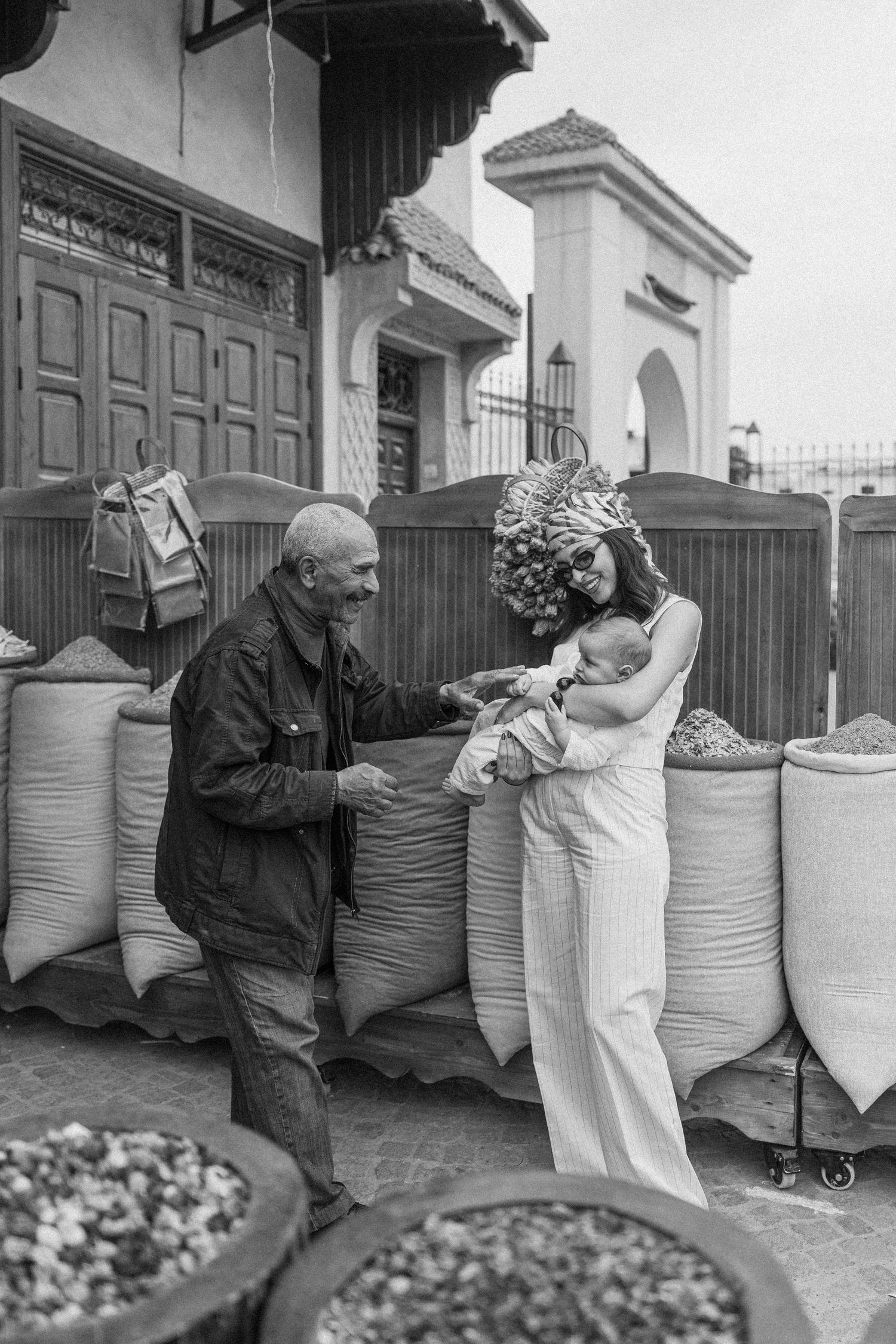 Black and white photo of a woman in white jumpsuit and floral headpiece holding a baby while interacting with an elderly man at a market.