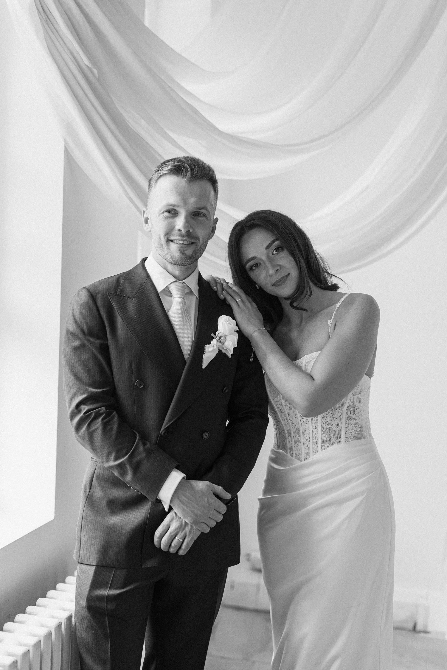 Black and white portrait of smiling groom in suit and bride in lace gown posing together at wedding.