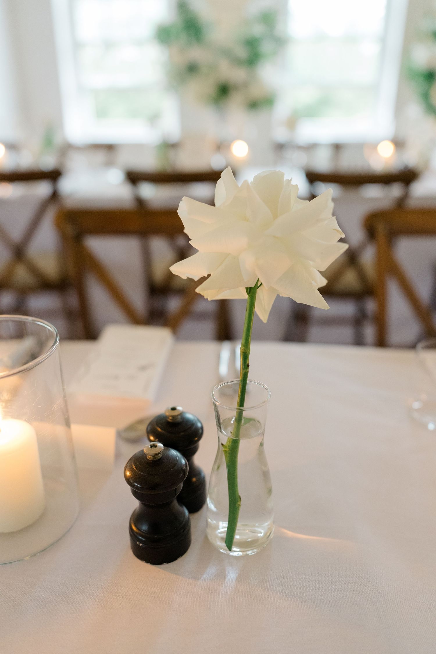 Single white narcissus flower in glass vase beside candle and salt pepper shakers on white tablecloth.