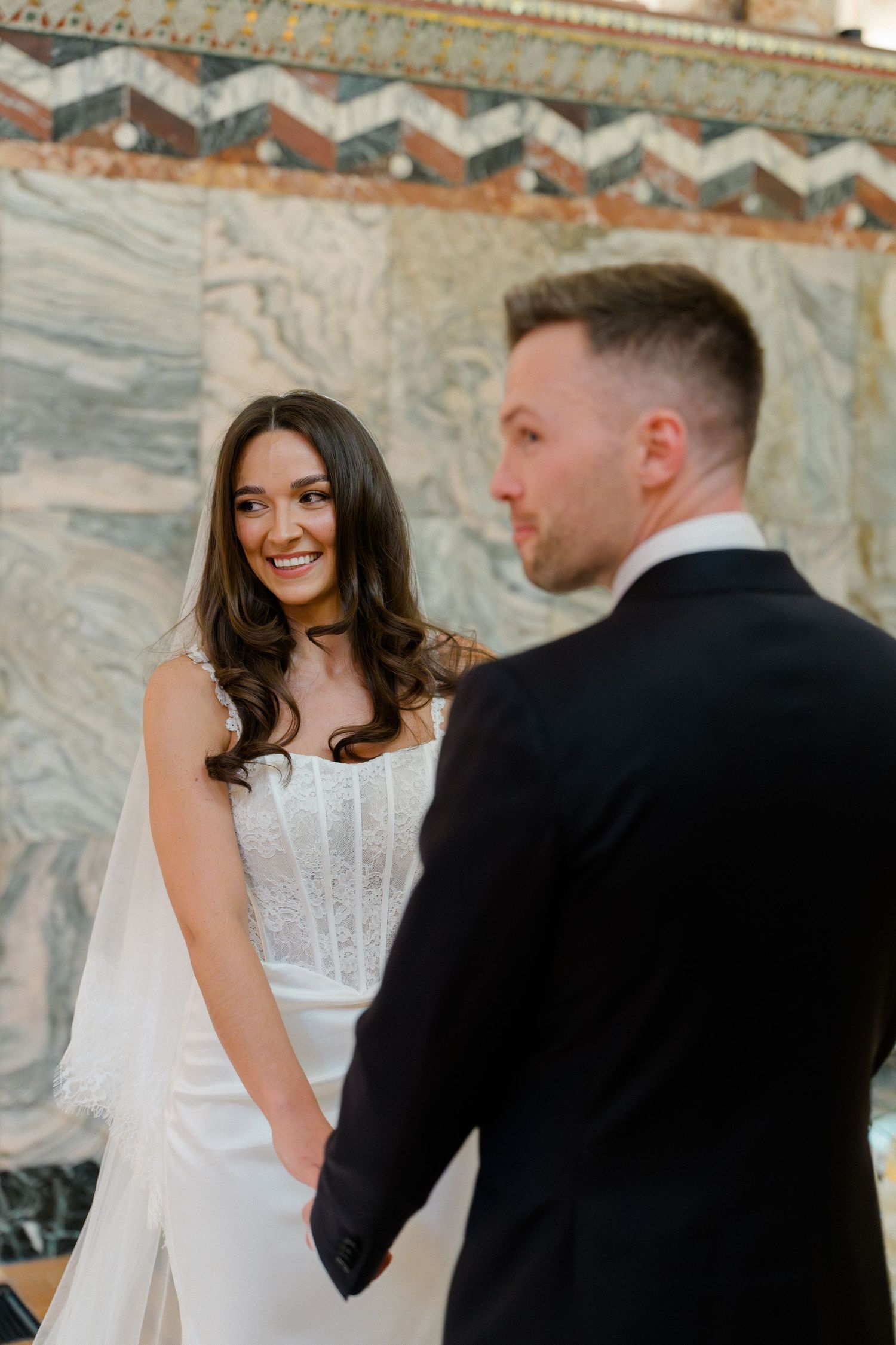 Smiling bride in white lace gown holds hands with groom in black suit during wedding ceremony.