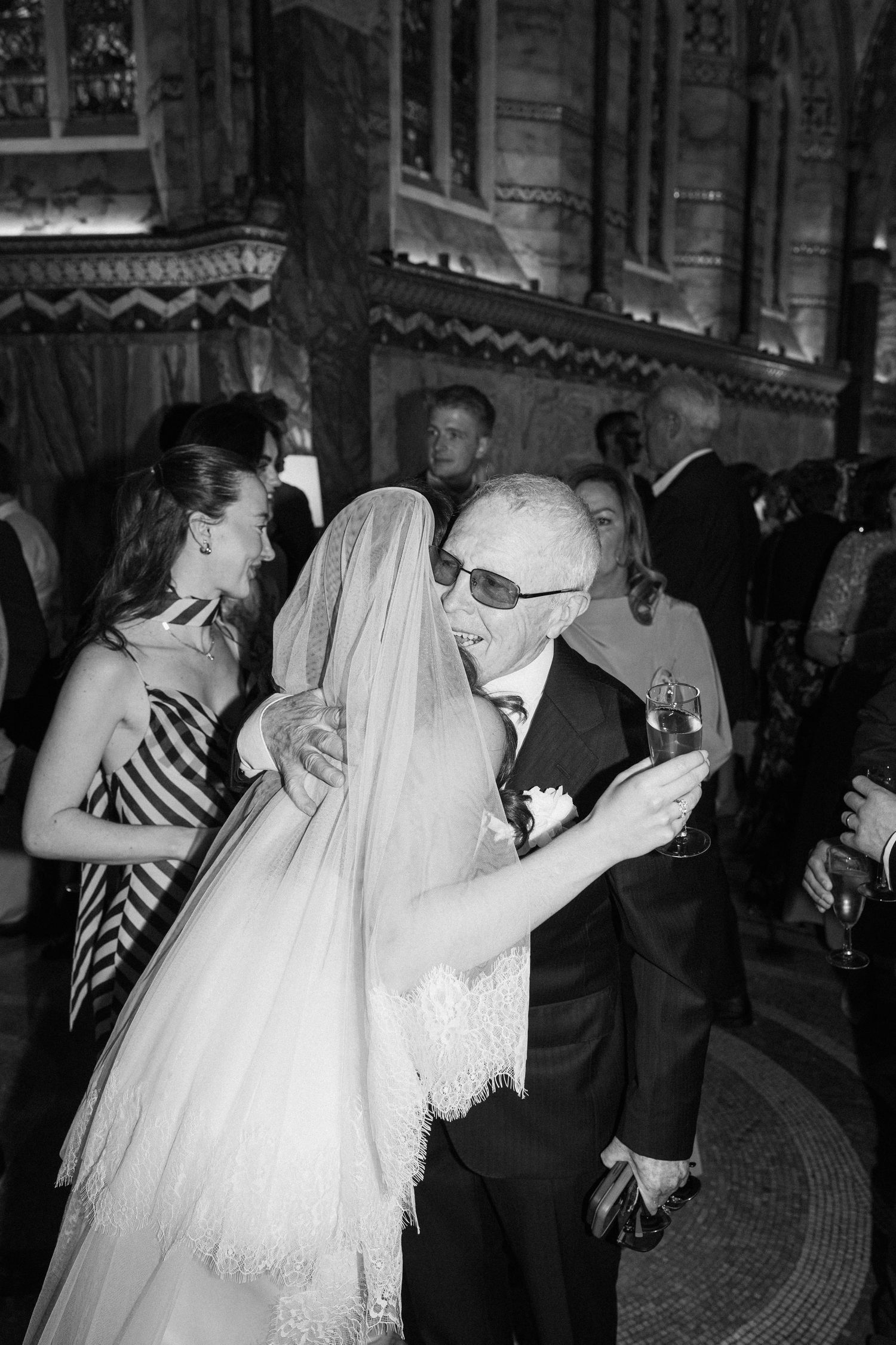 Black and white photo of a bride in veil embracing an elderly man in sunglasses at a wedding reception.
