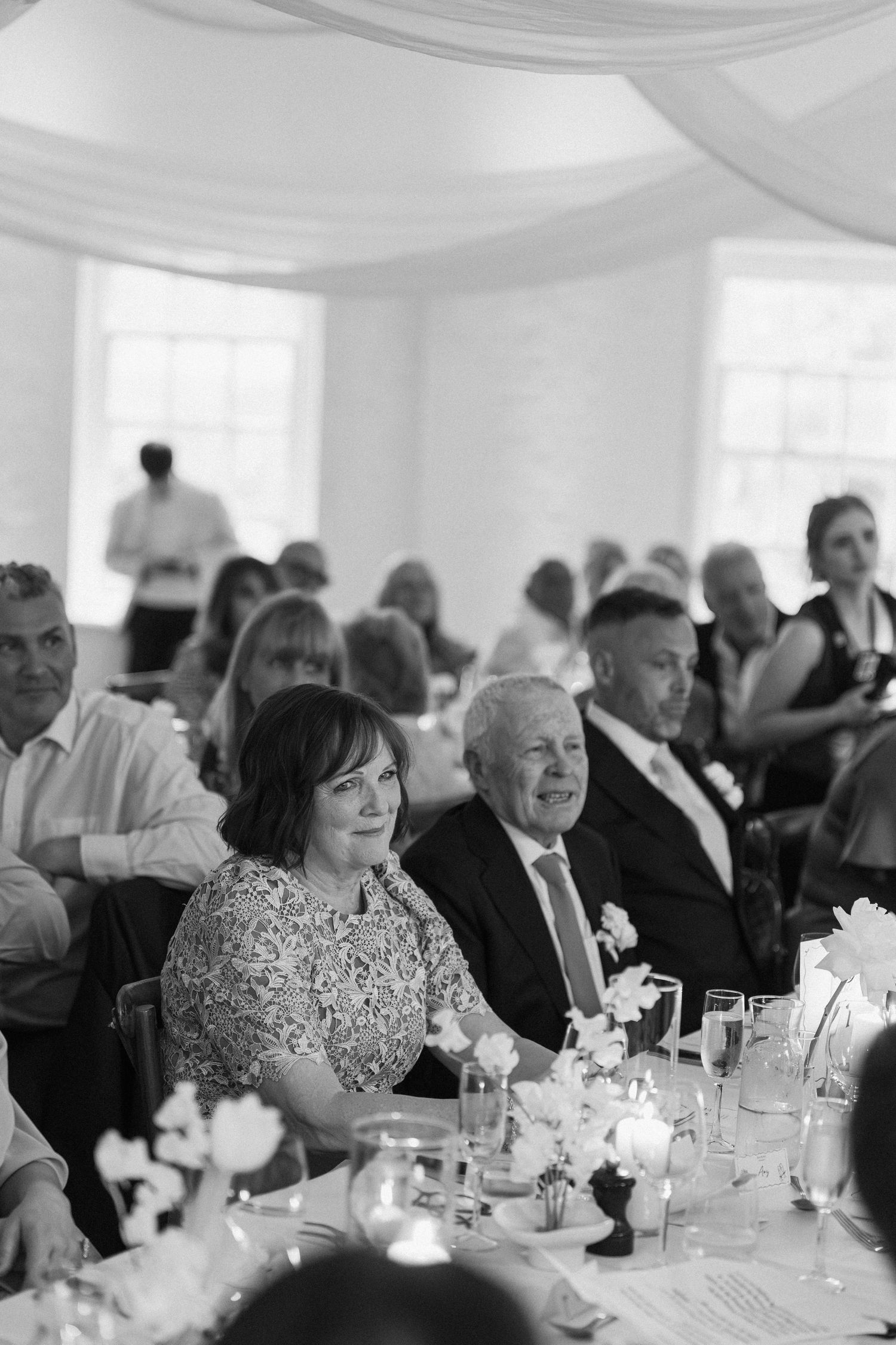 Wedding guests seated at reception tables smiling during speeches in beautifully decorated white venue.