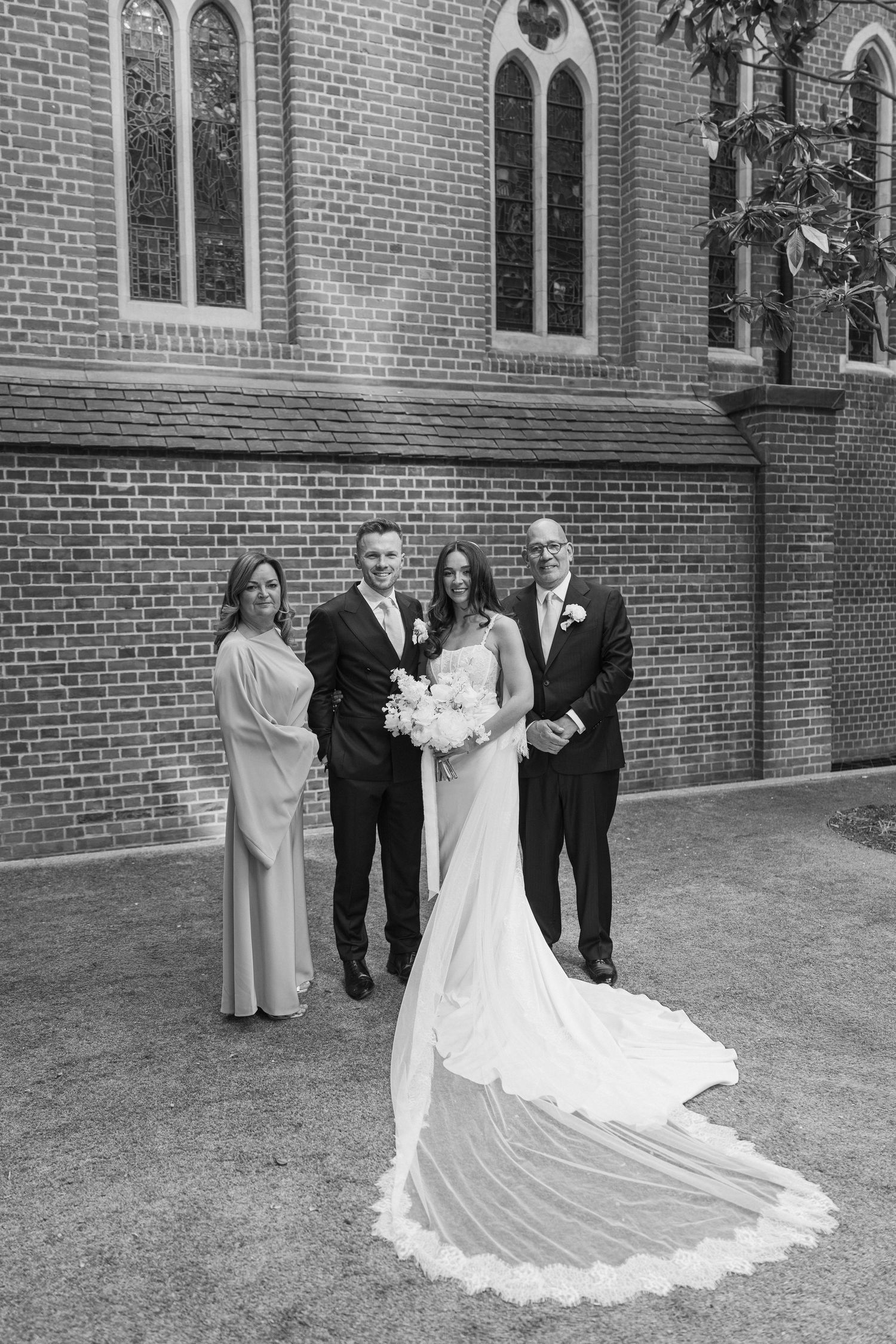 Black and white photo of a bride and groom posing with family outside a brick church after their wedding ceremony.
