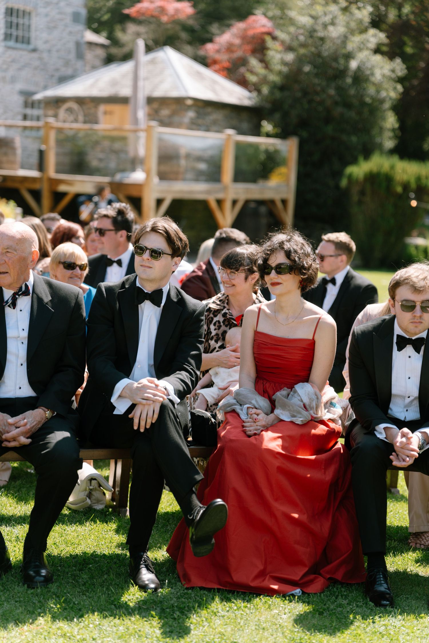 Wedding guests in formal attire and red dresses relax on grass during an outdoor ceremony.