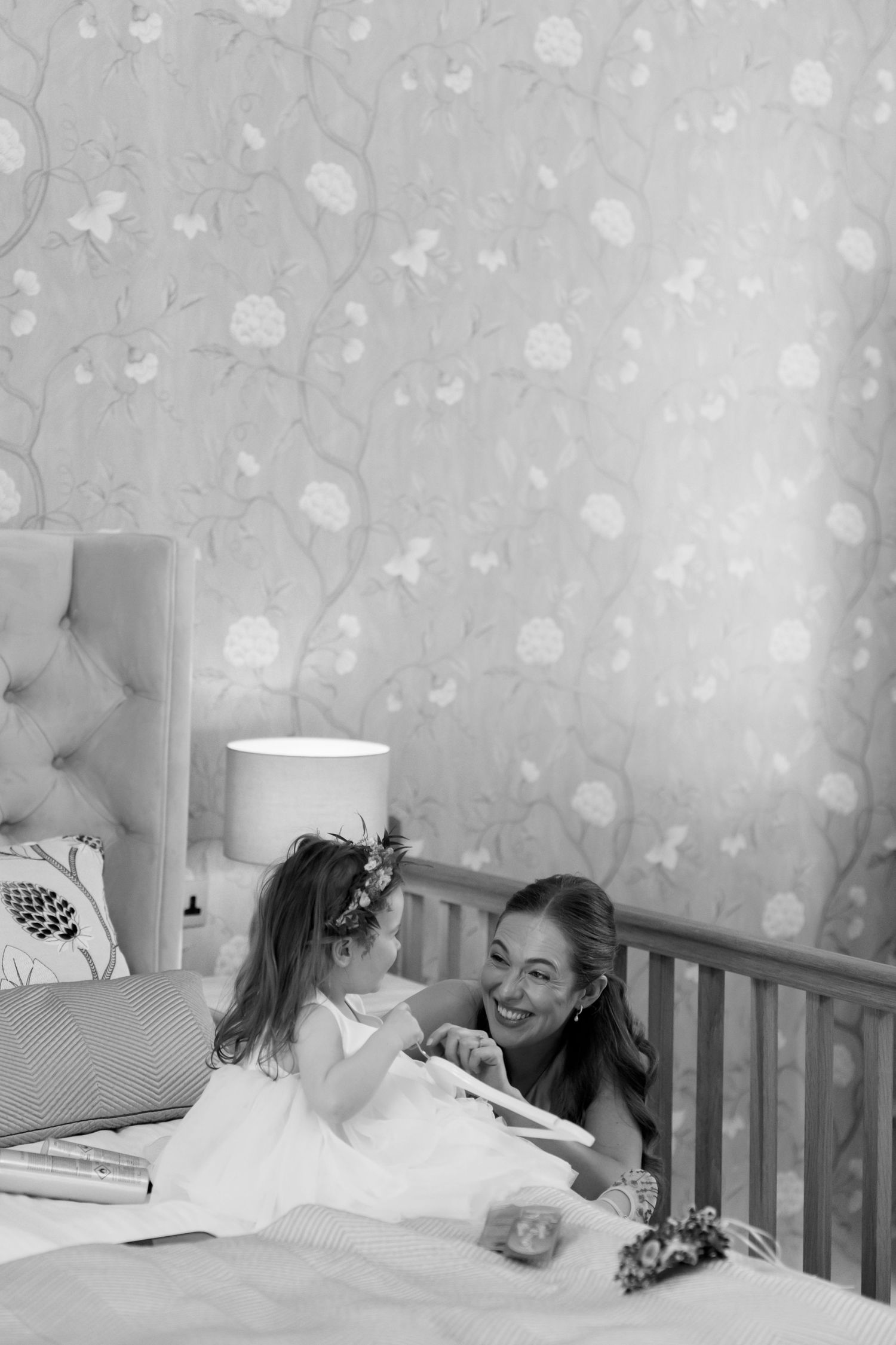 A tender black and white moment between a flower girl and bride on a bed with floral wallpaper backdrop.