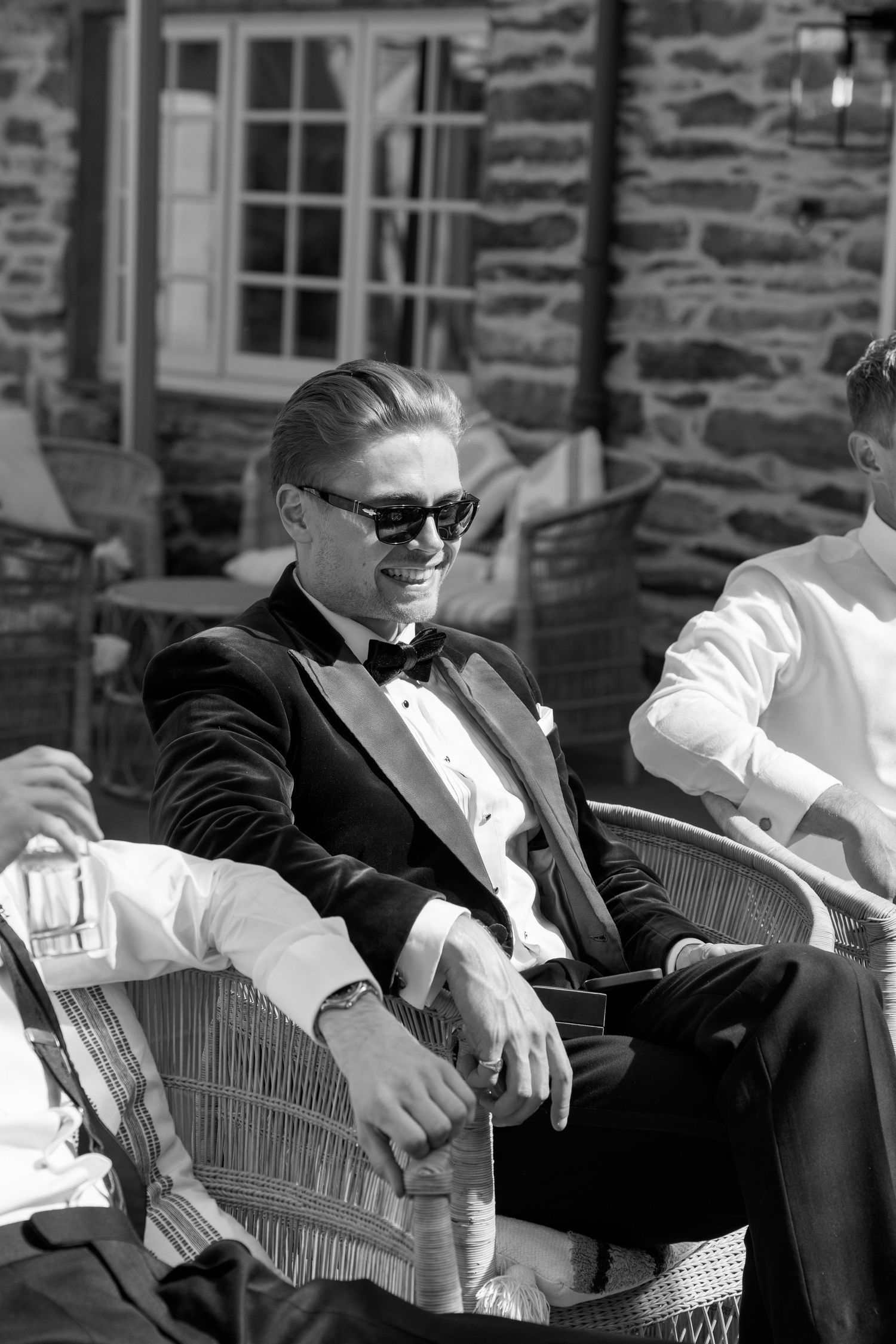Wedding party members in tuxedos lounging outdoors against stone building.