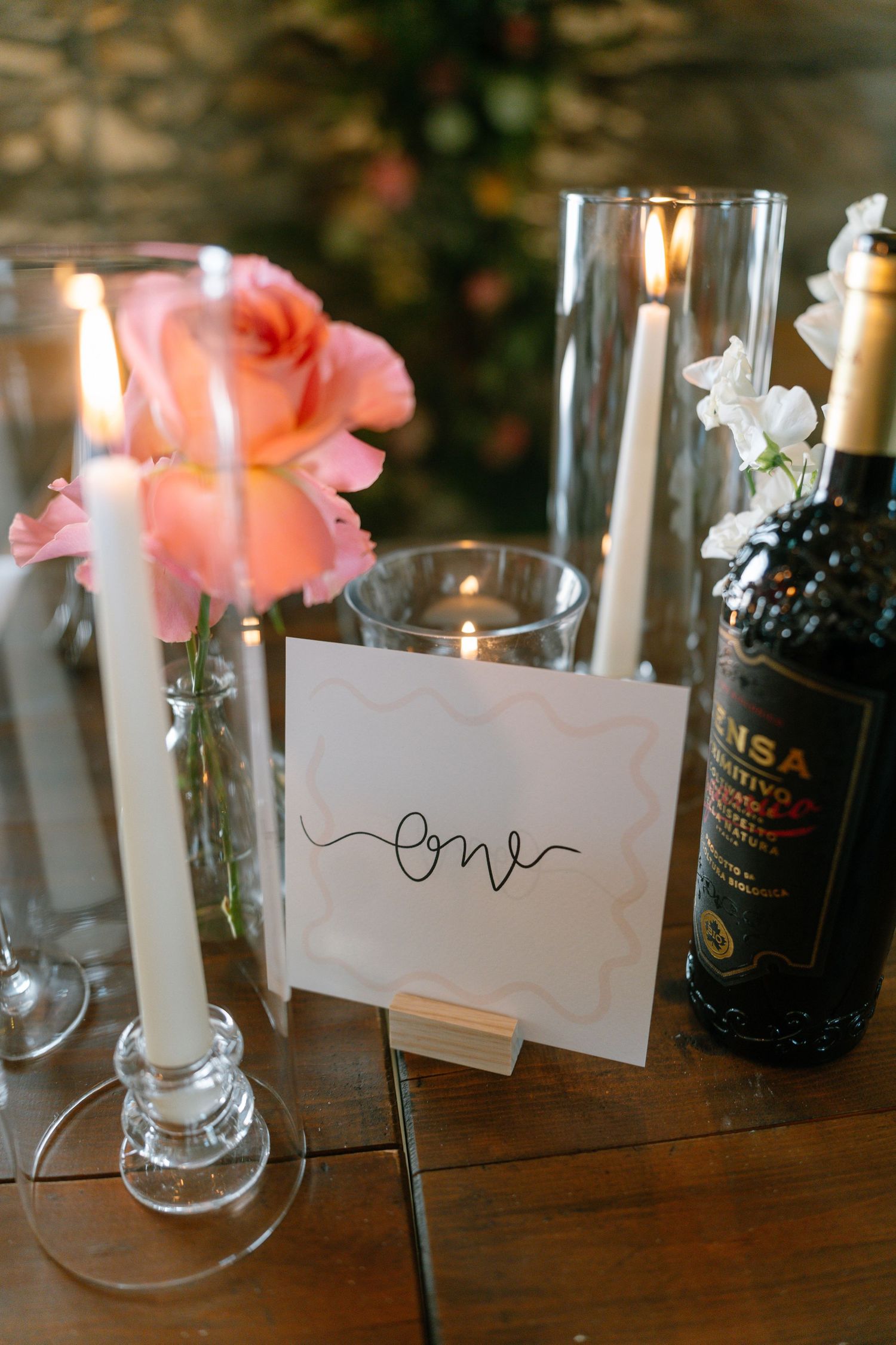 Wedding table decor featuring pink flowers, wine bottles, and handwritten place cards on dark wood.