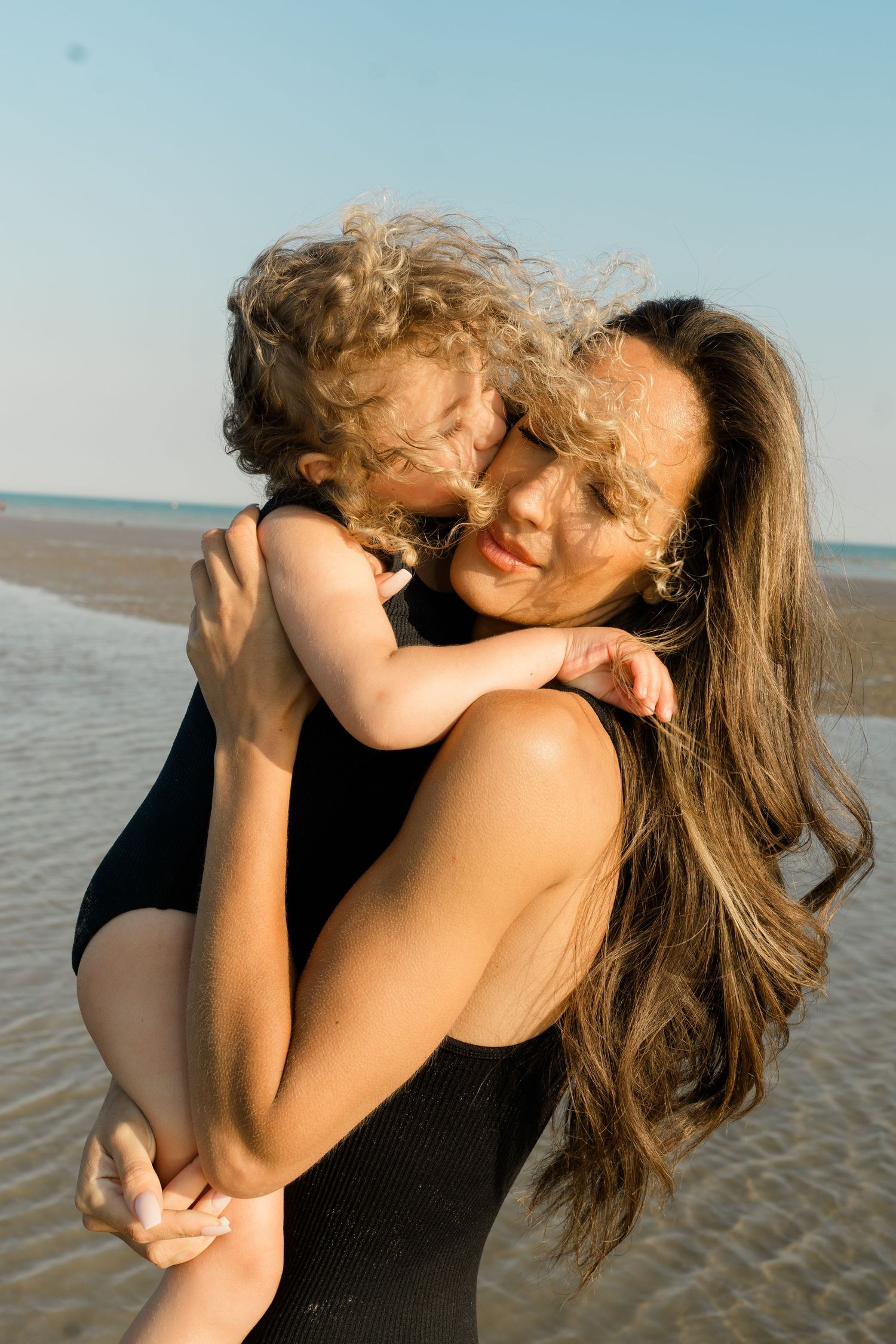 Two people in black swimsuits embrace joyfully on a sunny beach.