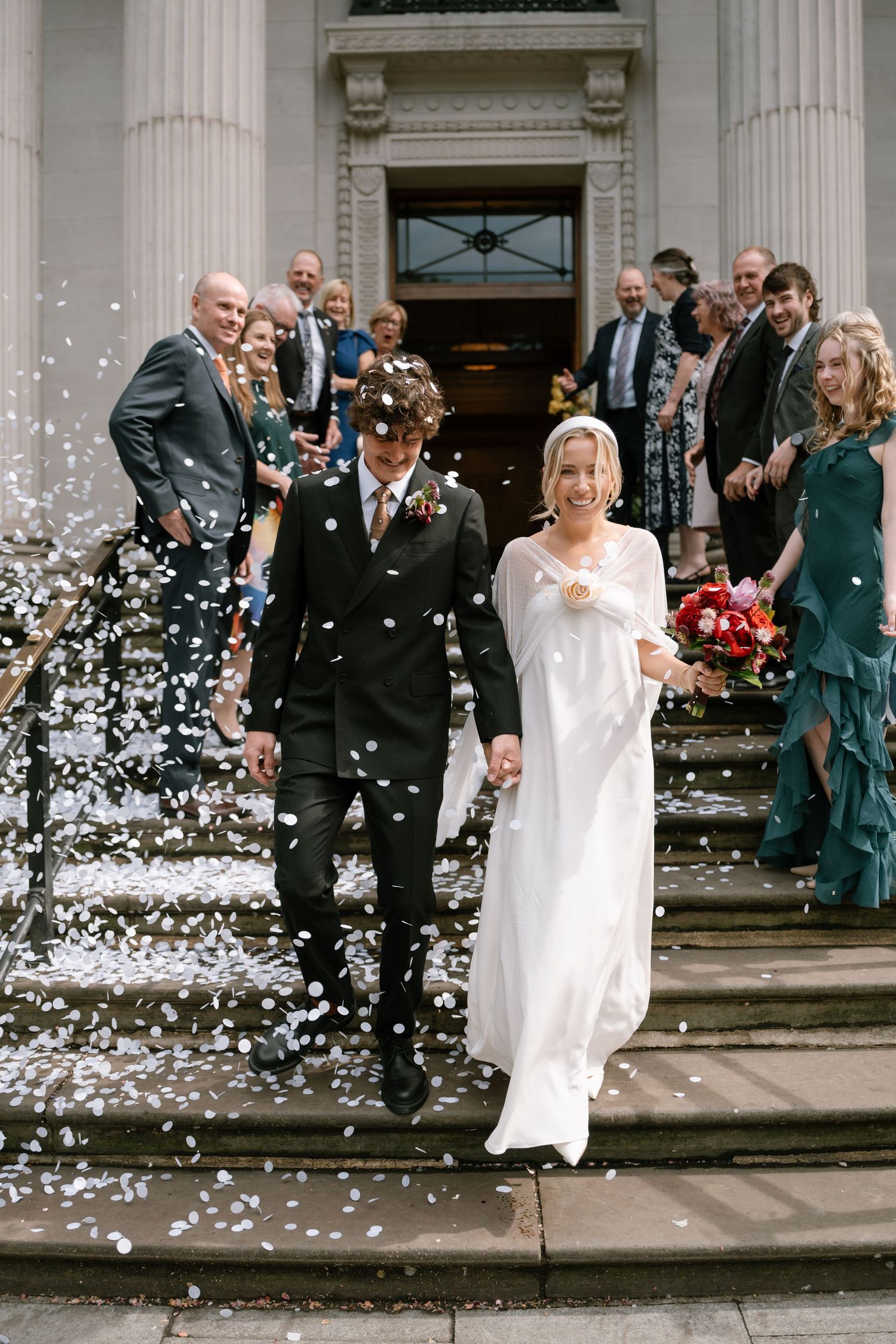 Wedding party celebrates with confetti toss on steps of columned building entrance.