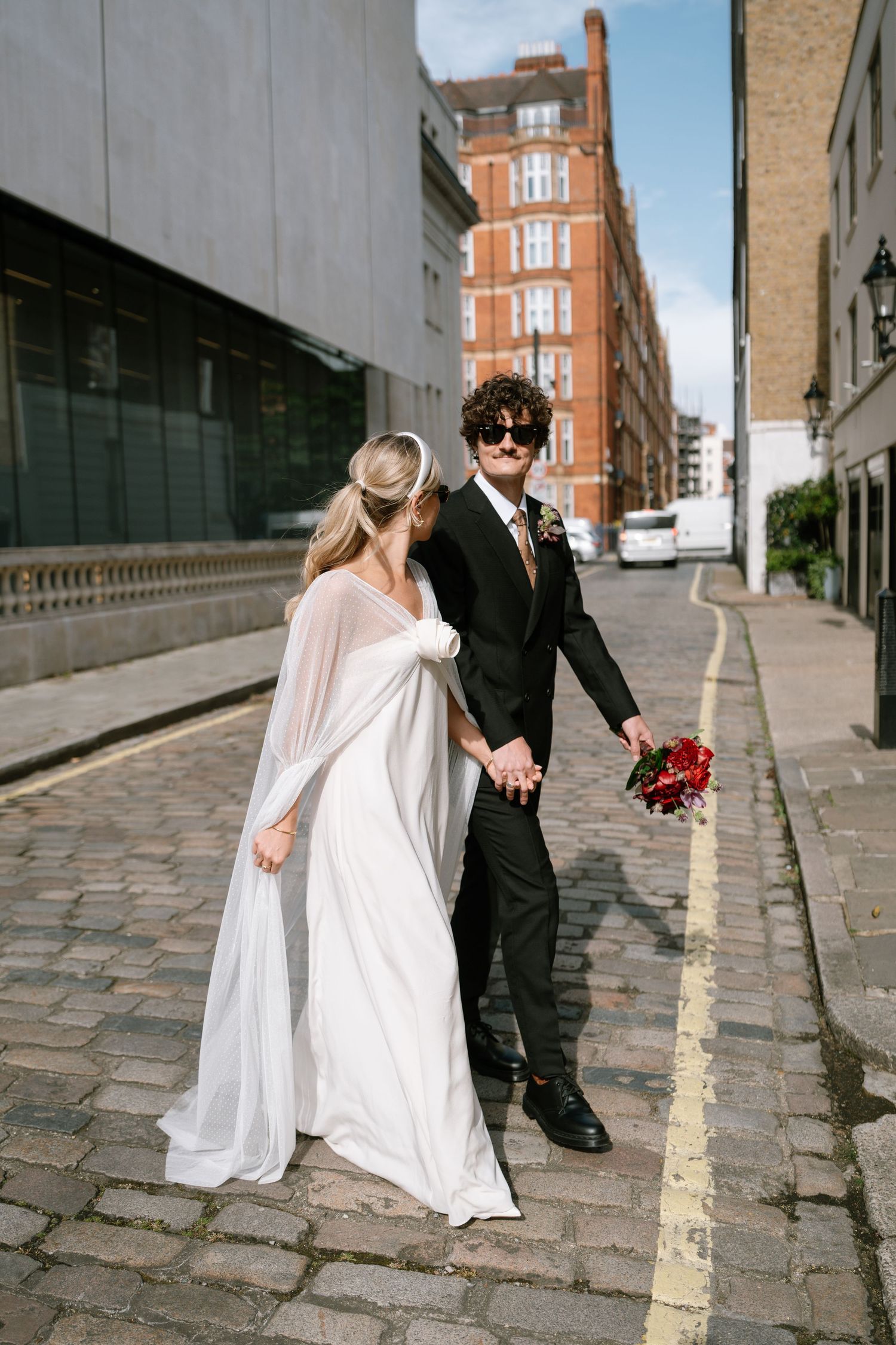 A couple in wedding attire poses on a cobblestone street in front of a brick building.