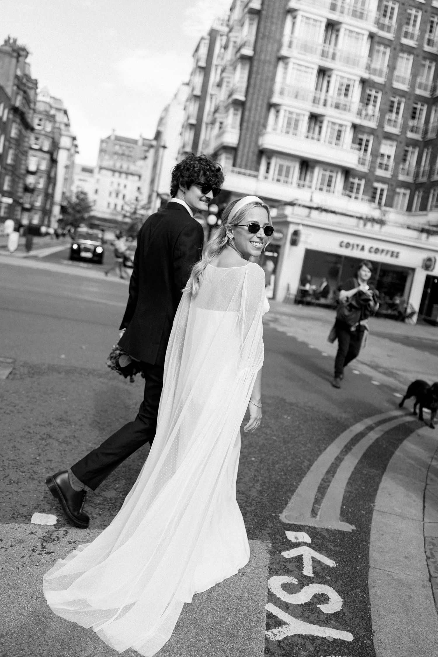 A couple walks across street markings in black and white with classic architecture in the background.