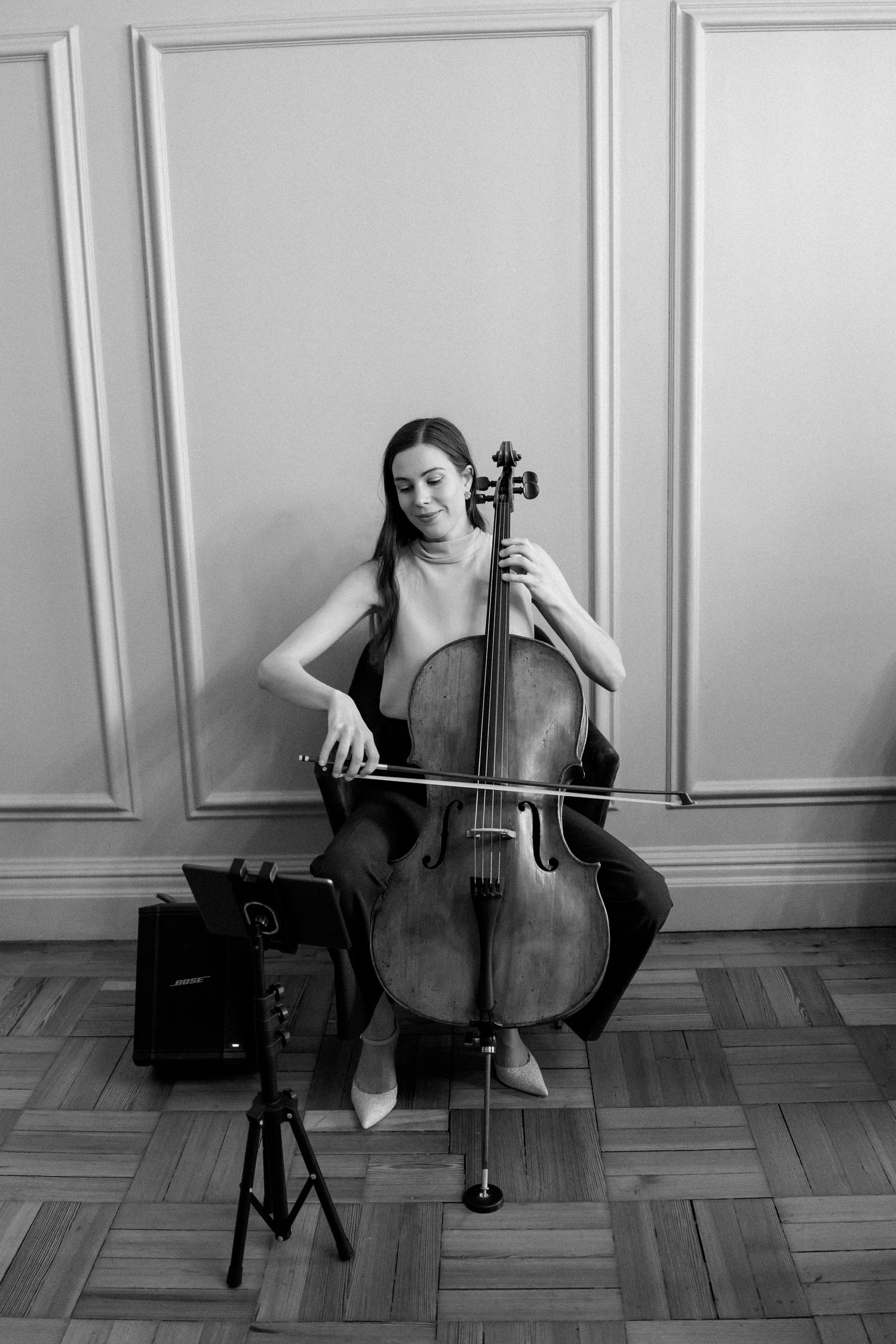 Black and white photo of a cellist performing in an elegant room with paneled walls.
