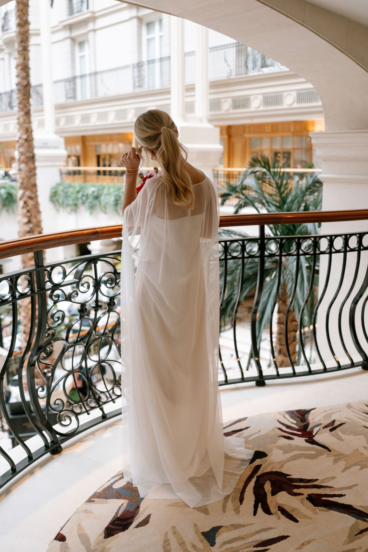 Bride in white dress standing on balcony with ornate iron railings and palm trees.