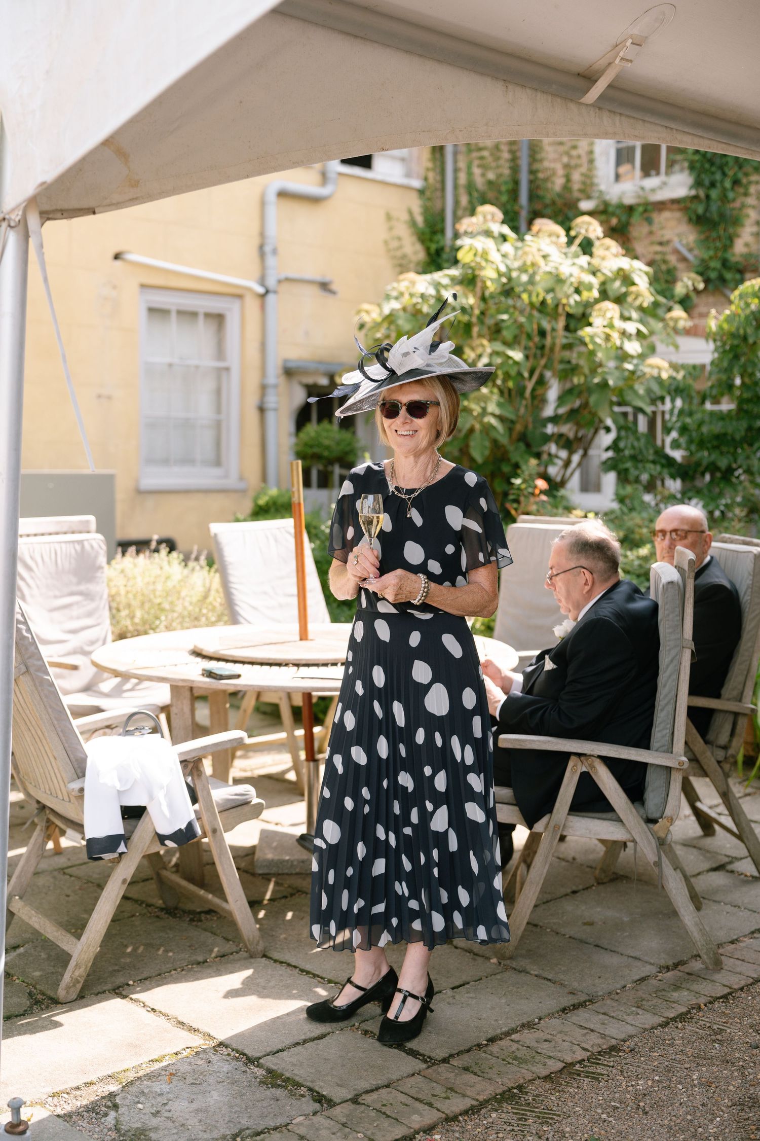 A person in a polka dot dress and sun hat sits on a wooden deck chair in an outdoor setting.