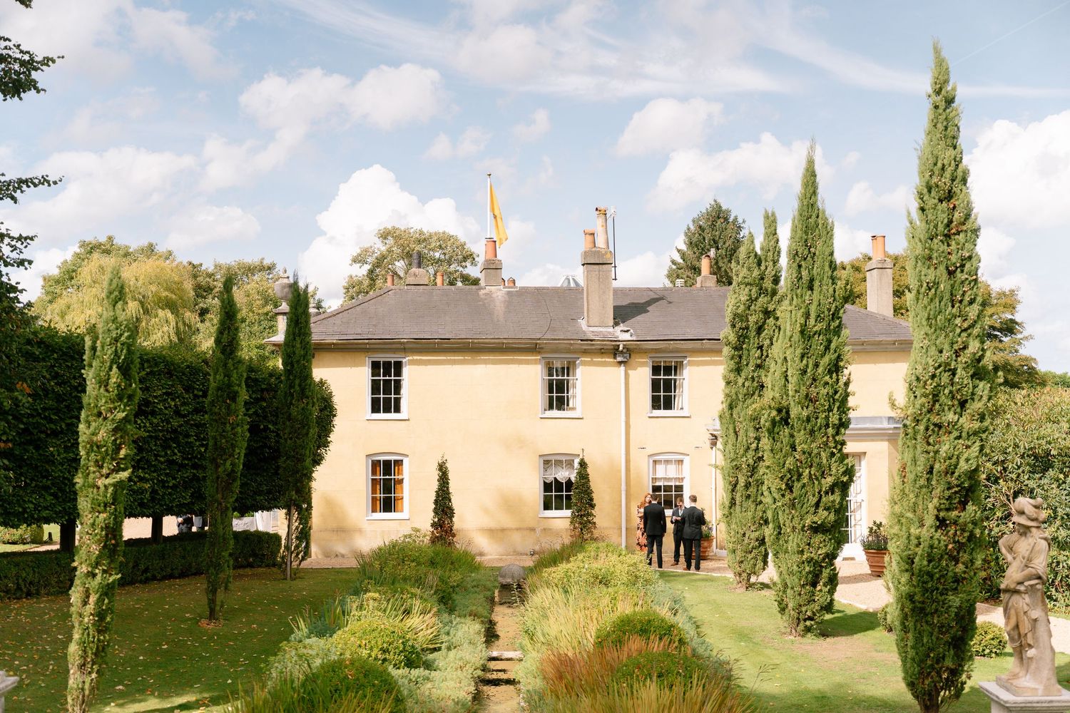 Exterior view of cream-colored country estate with cypress trees and landscaped grounds.