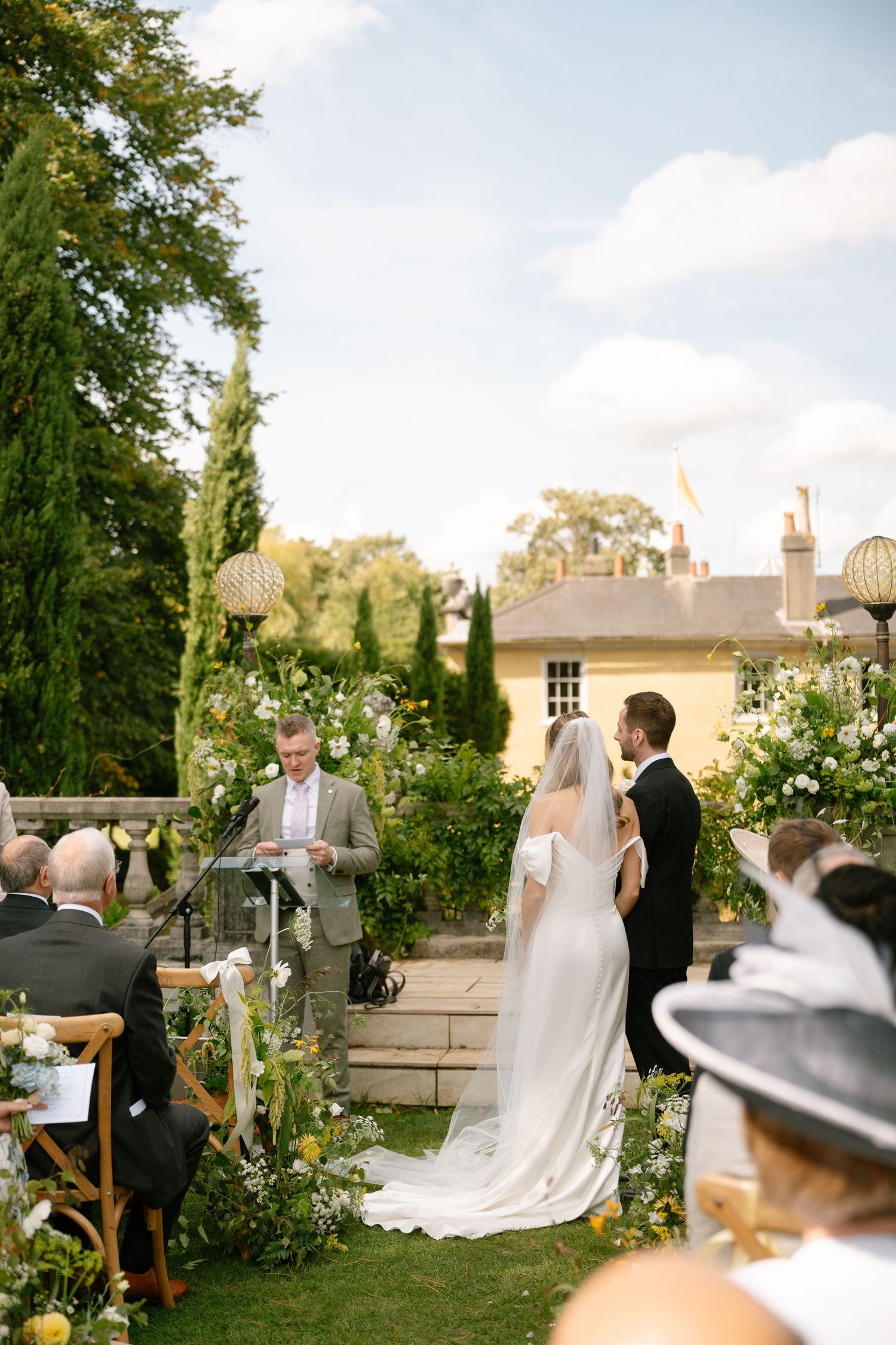 Outdoor wedding ceremony takes place in a beautiful garden setting with white chairs and floral decorations.