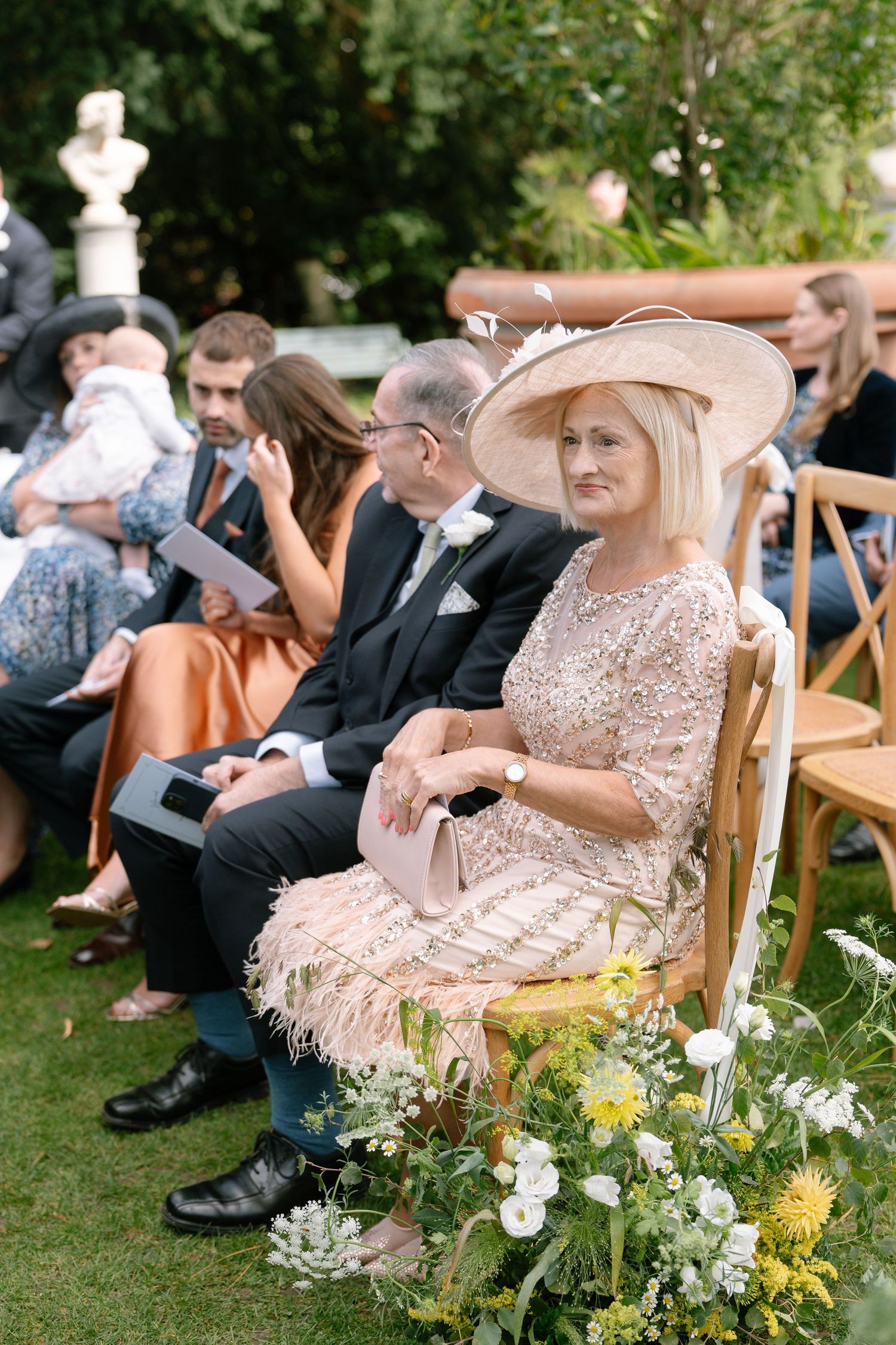 Wedding guests gather on a lawn with wooden chairs and floral decorations at an outdoor garden ceremony.