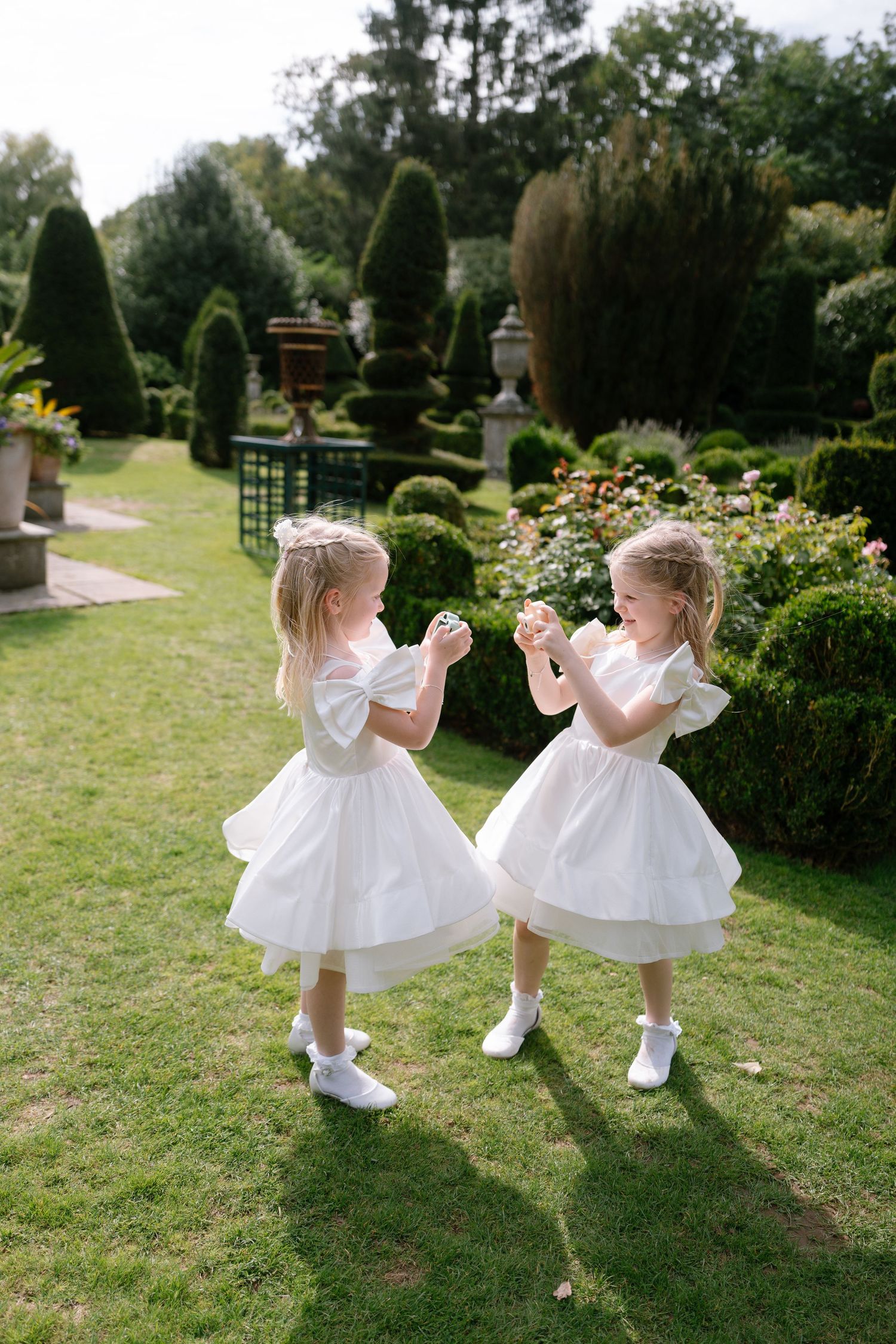 Two young flower girls in white dresses dancing playfully on a manicured lawn.