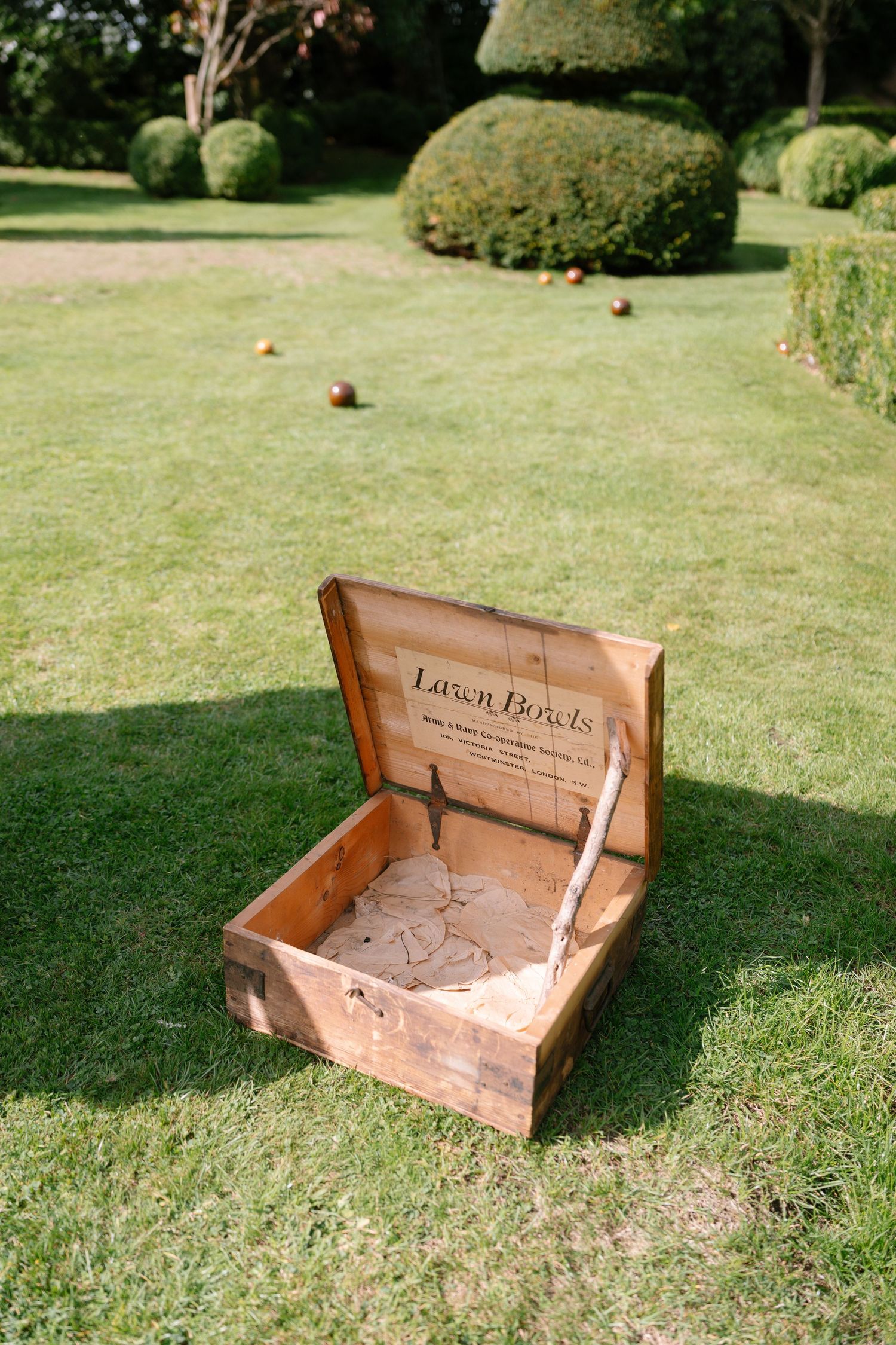 Wooden lawn petanque game box placed on manicured grass with topiary bushes in background.