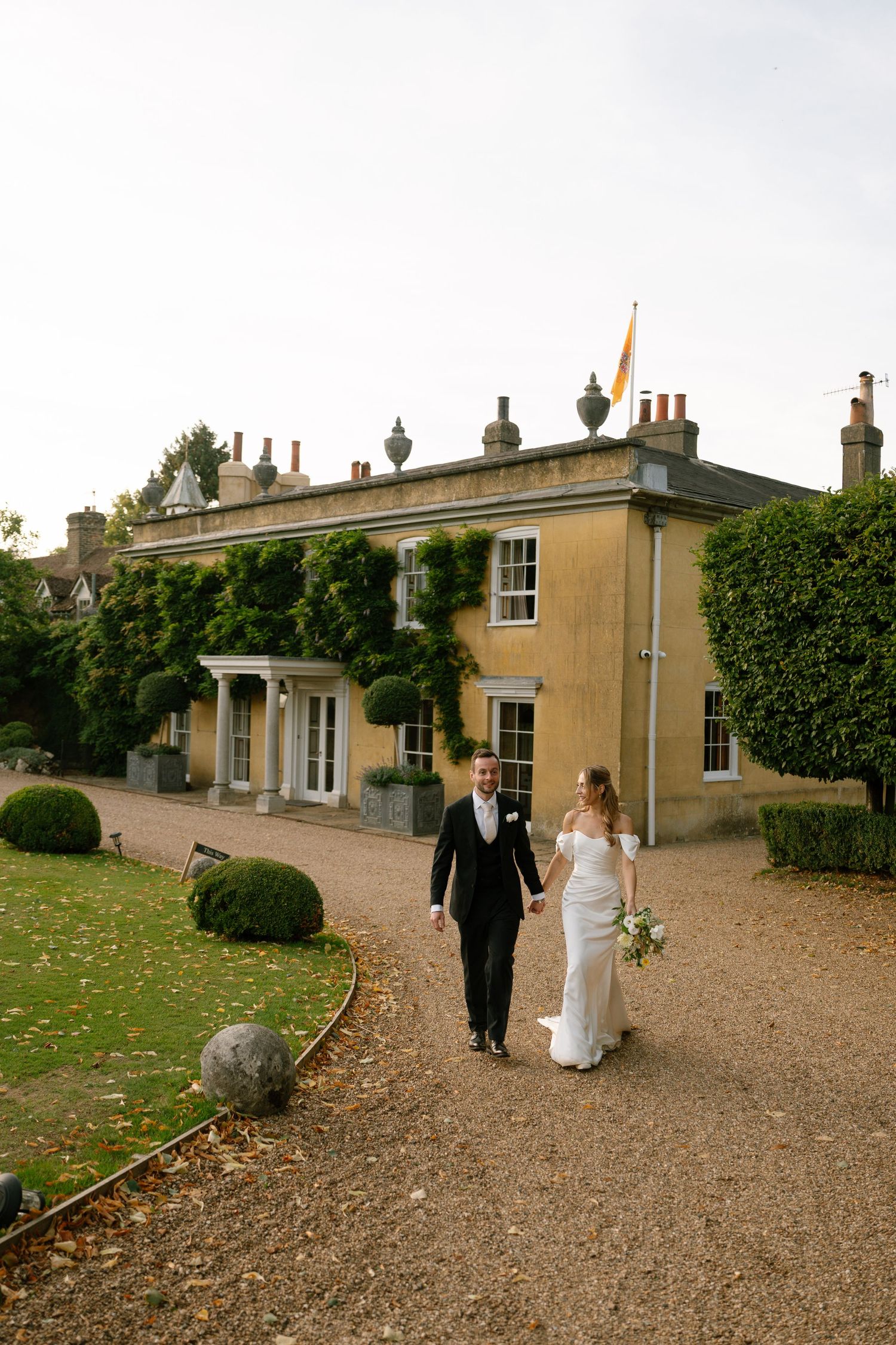 A wedding couple walks hand in hand along a gravel path toward a stately manor house.
