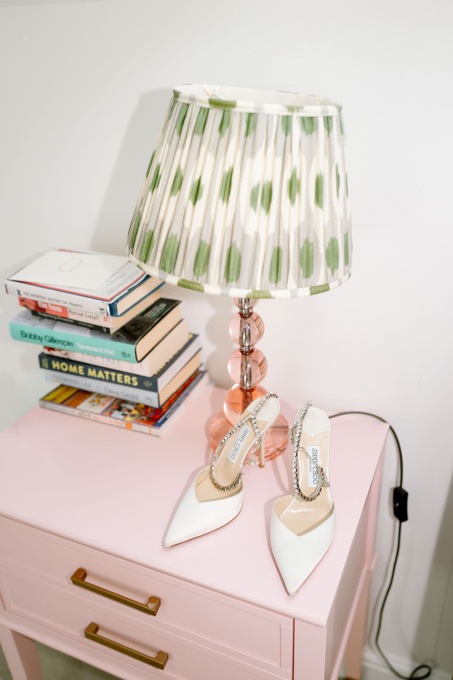 Pink bedside table with vintage lamp, white shoes, and stacked books creating a romantic wedding detail.