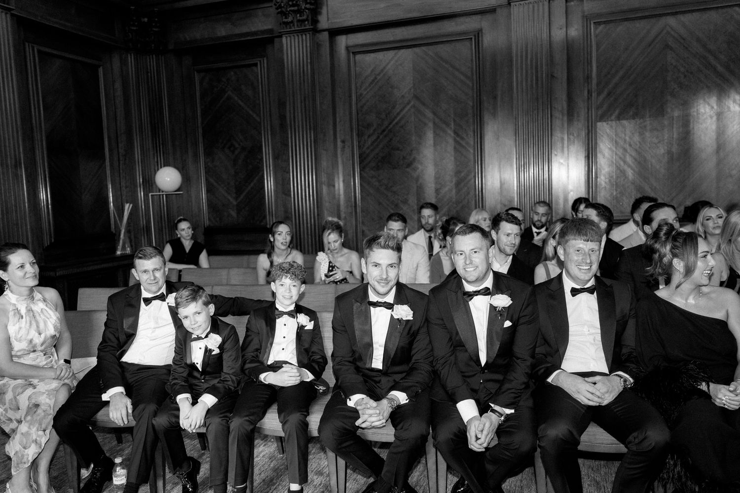 Wedding guests in formal attire seated in rows of wooden chairs during ceremony.