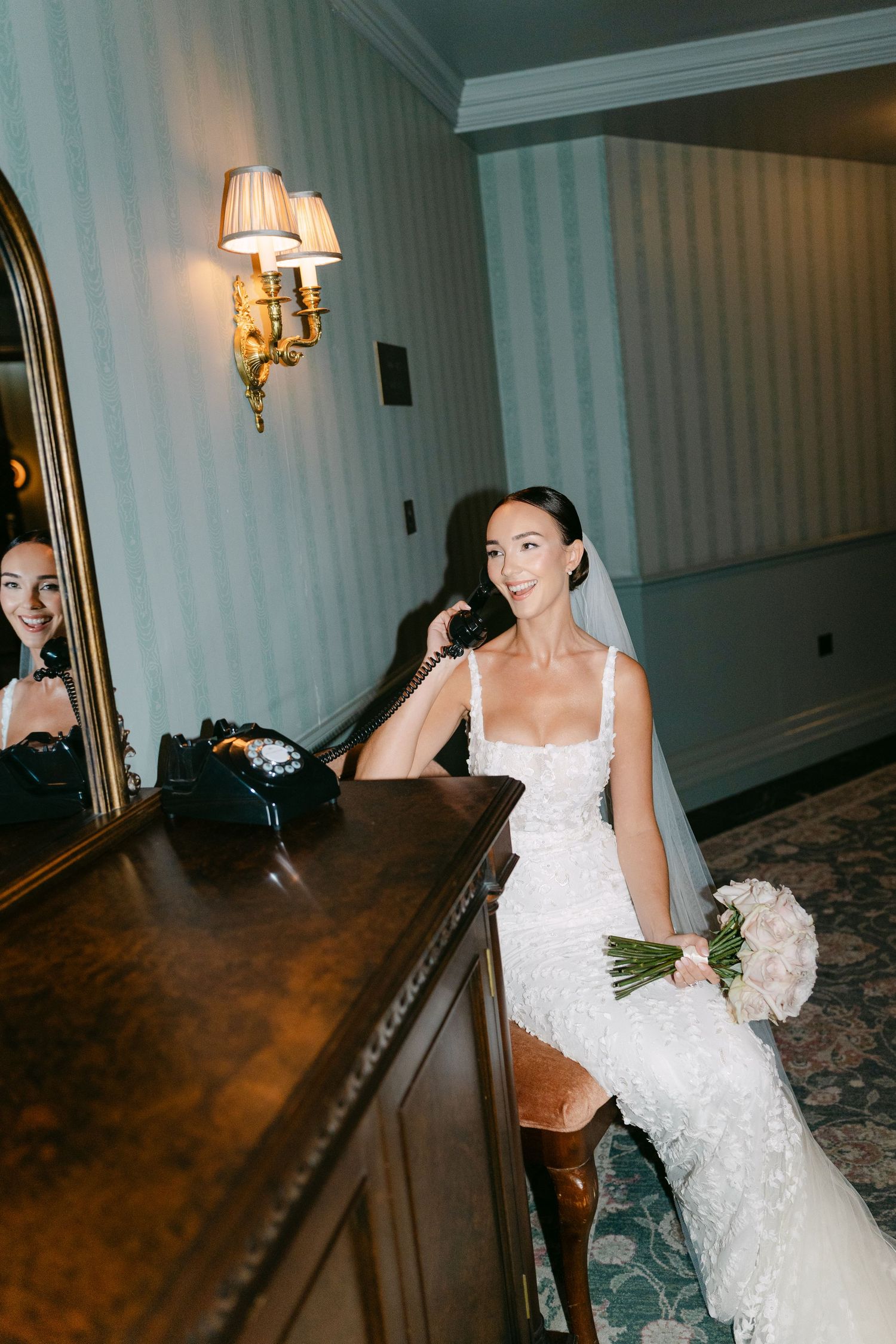Elegant bride in lace gown poses against dark wood furniture in classic interior setting.