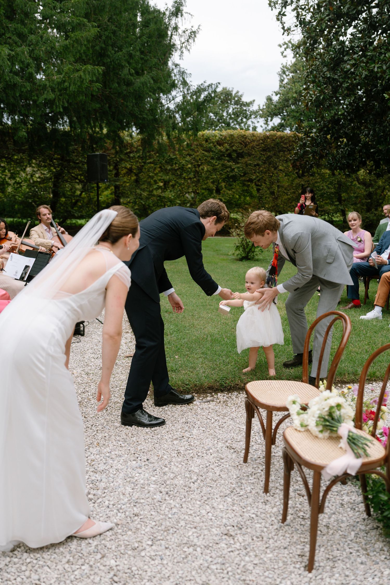 Wedding party members lean playfully on wooden chairs during a photo session on a gravel path.