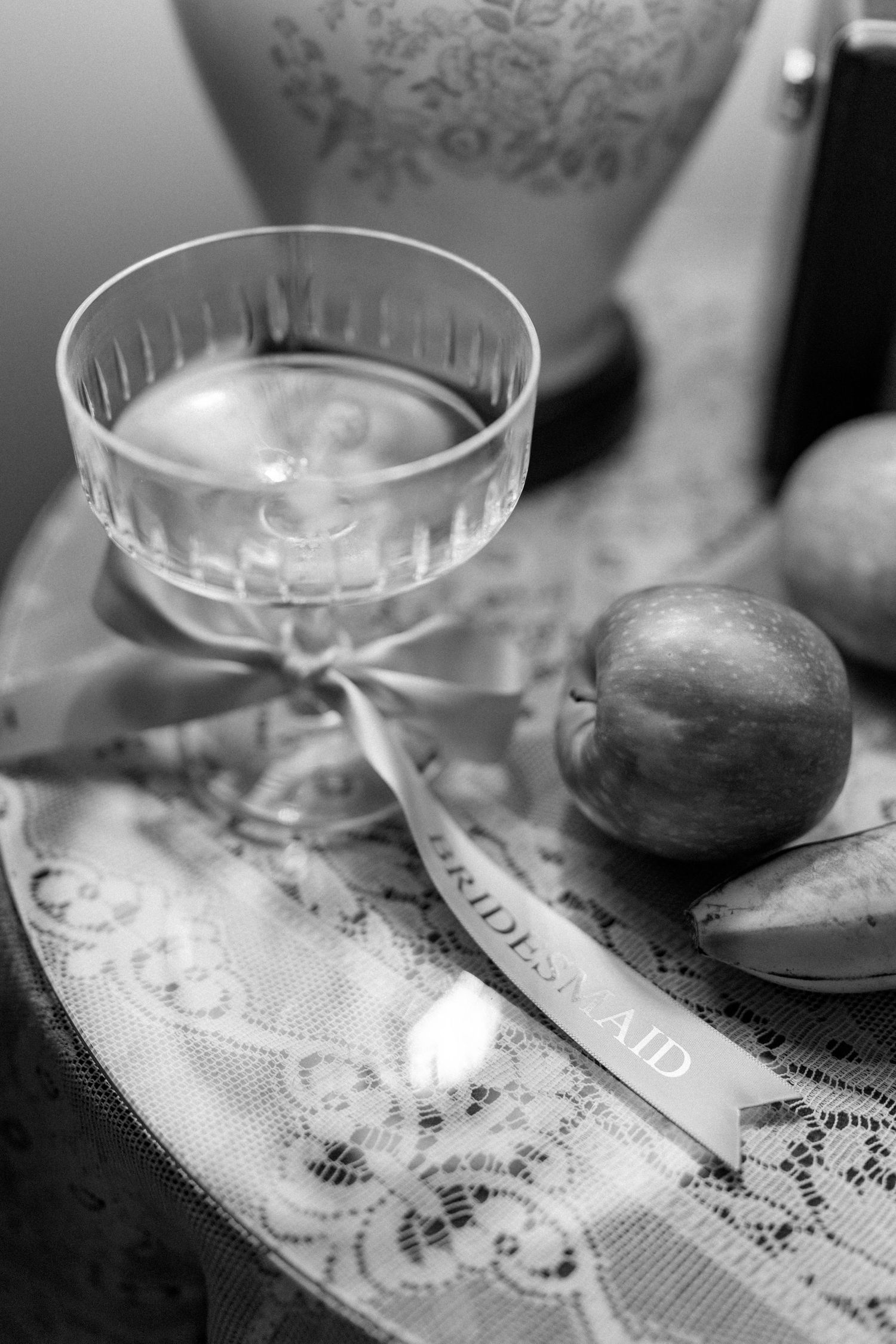 A vintage cocktail glass and fruit sit on a lace tablecloth in black and white.