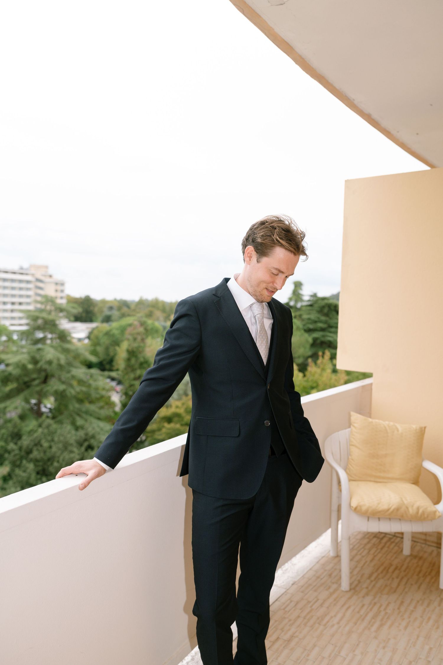 A person in a black suit stands on a balcony overlooking trees and buildings in the distance.