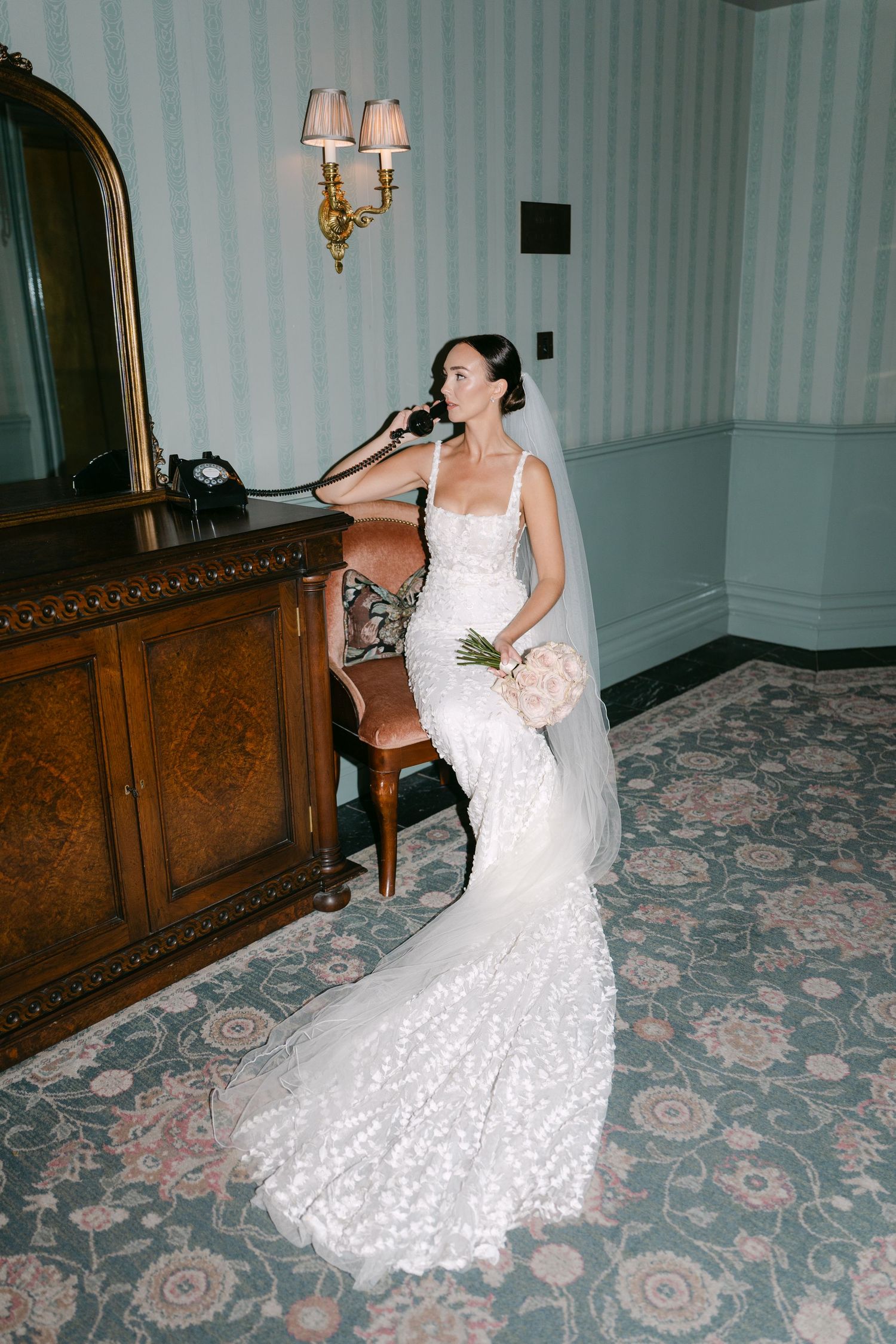 Bride in fitted lace gown poses elegantly in vintage-styled room with wall sconces.