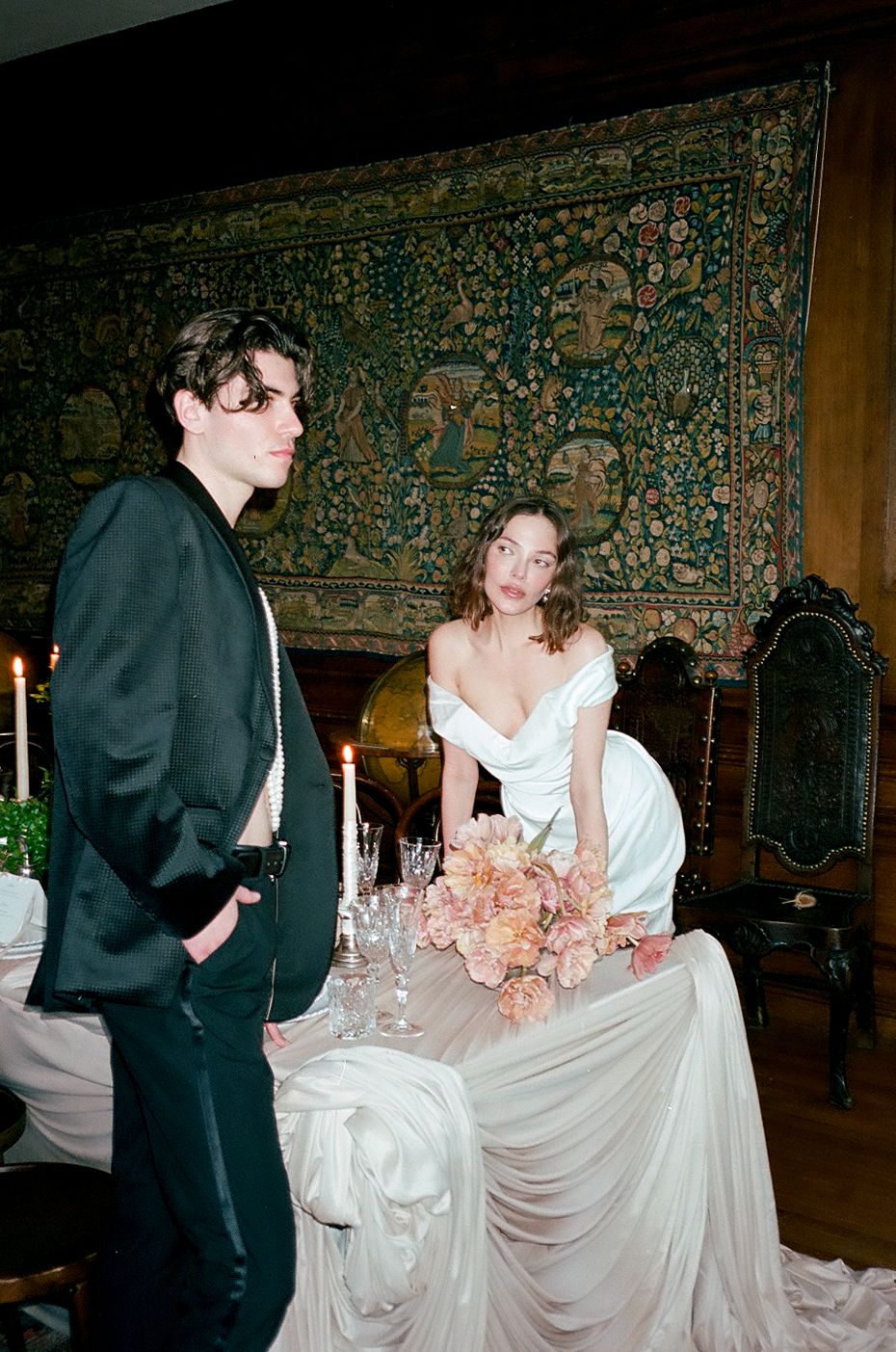 A bride in a white off-shoulder gown leans on a draped table with pink flowers beside a man in black suit.