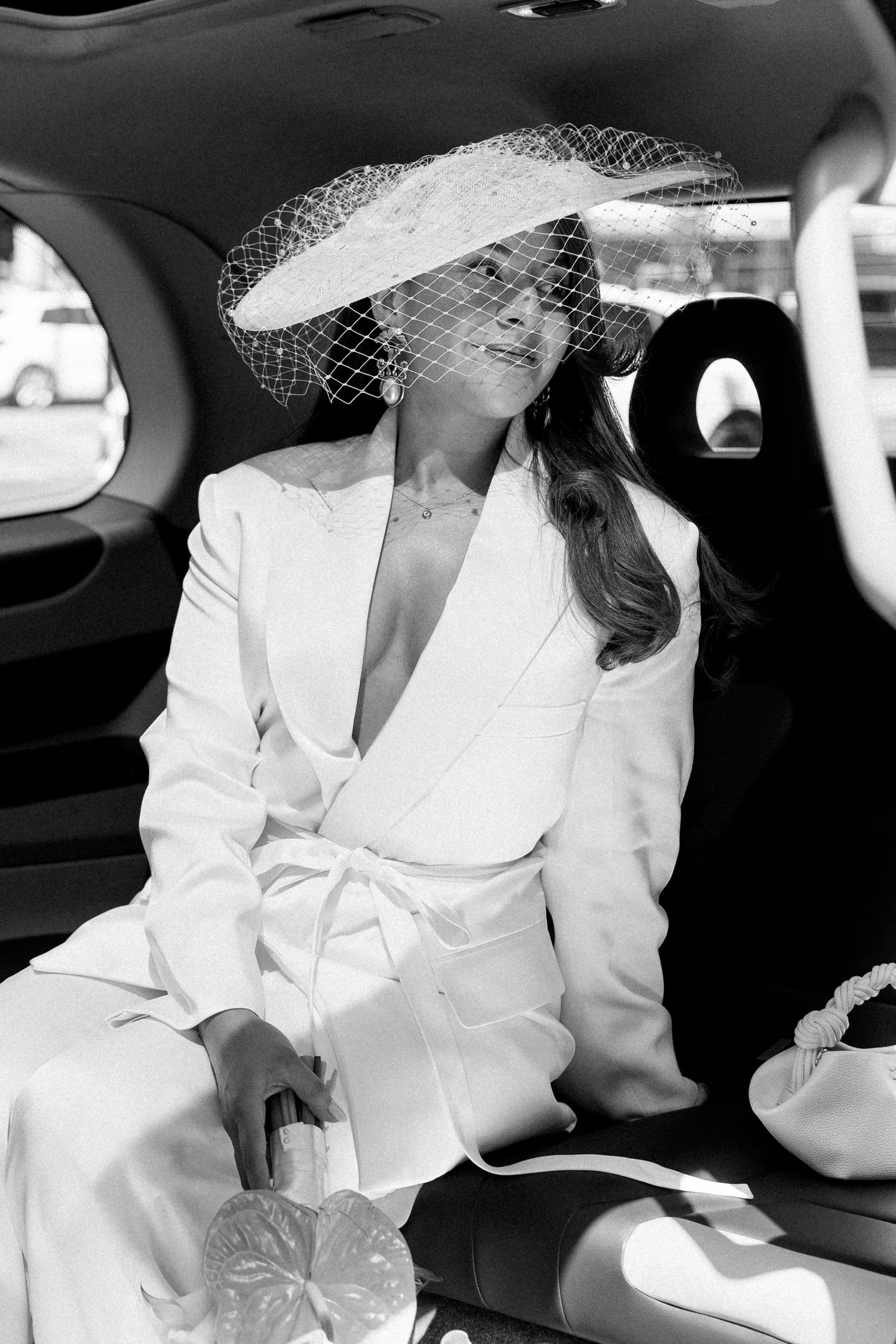 Elegant bride in white belted blazer suit and wide-brim veil hat seated in car, black and white photo.