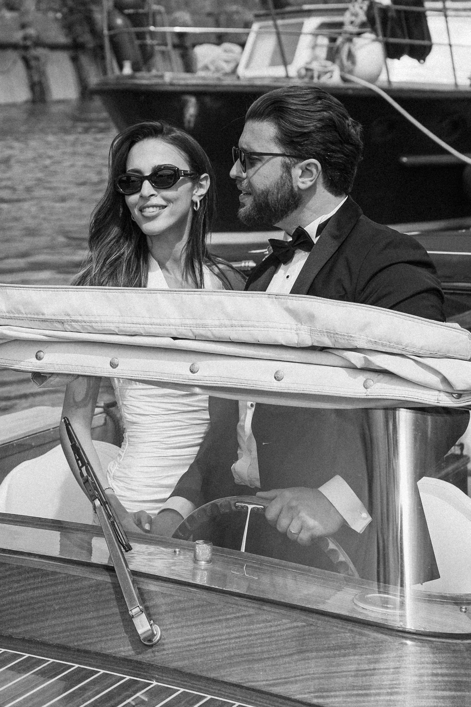 Elegant couple in formal attire riding a classic boat, woman in white dress with sunglasses, man in tuxedo.