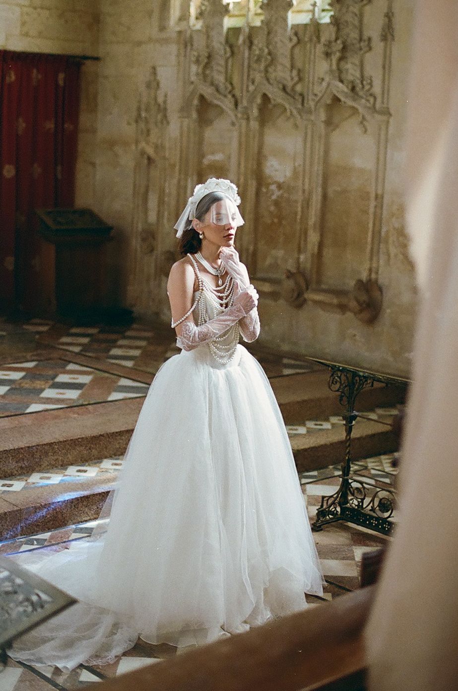 A bride in a voluminous white ball gown and pearls poses gracefully in an ornate church interior.