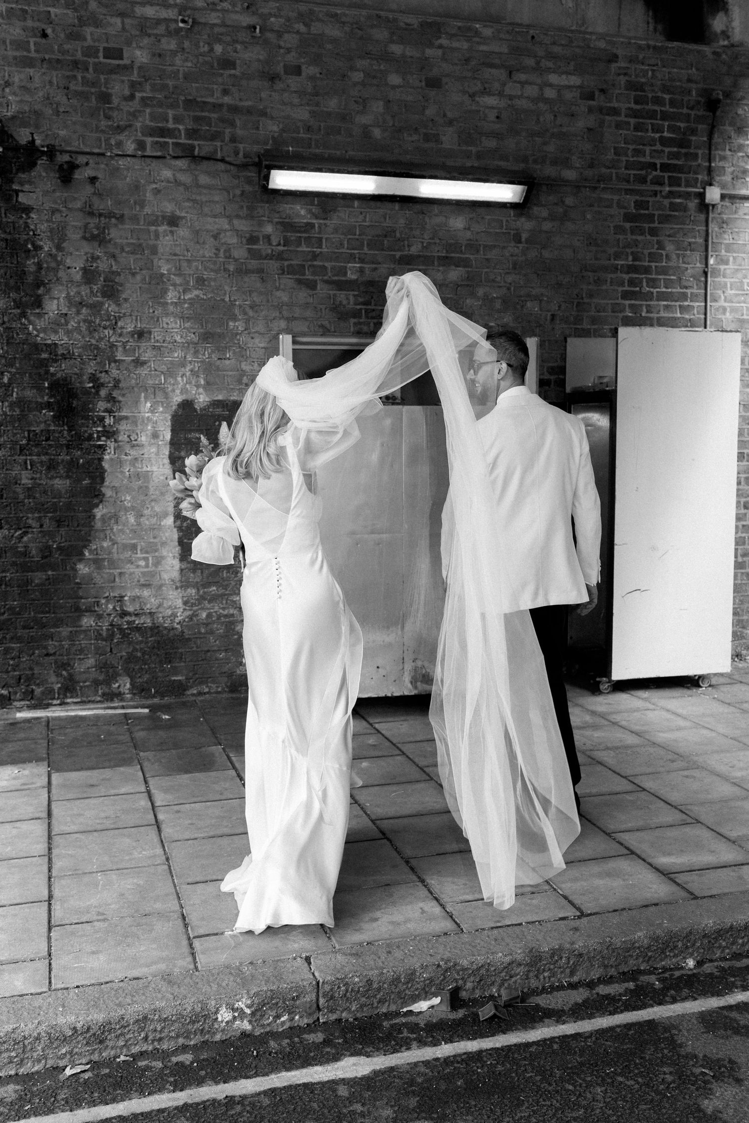 A black and white photo of a couple in white outfits dancing together on a brick-lined street.
