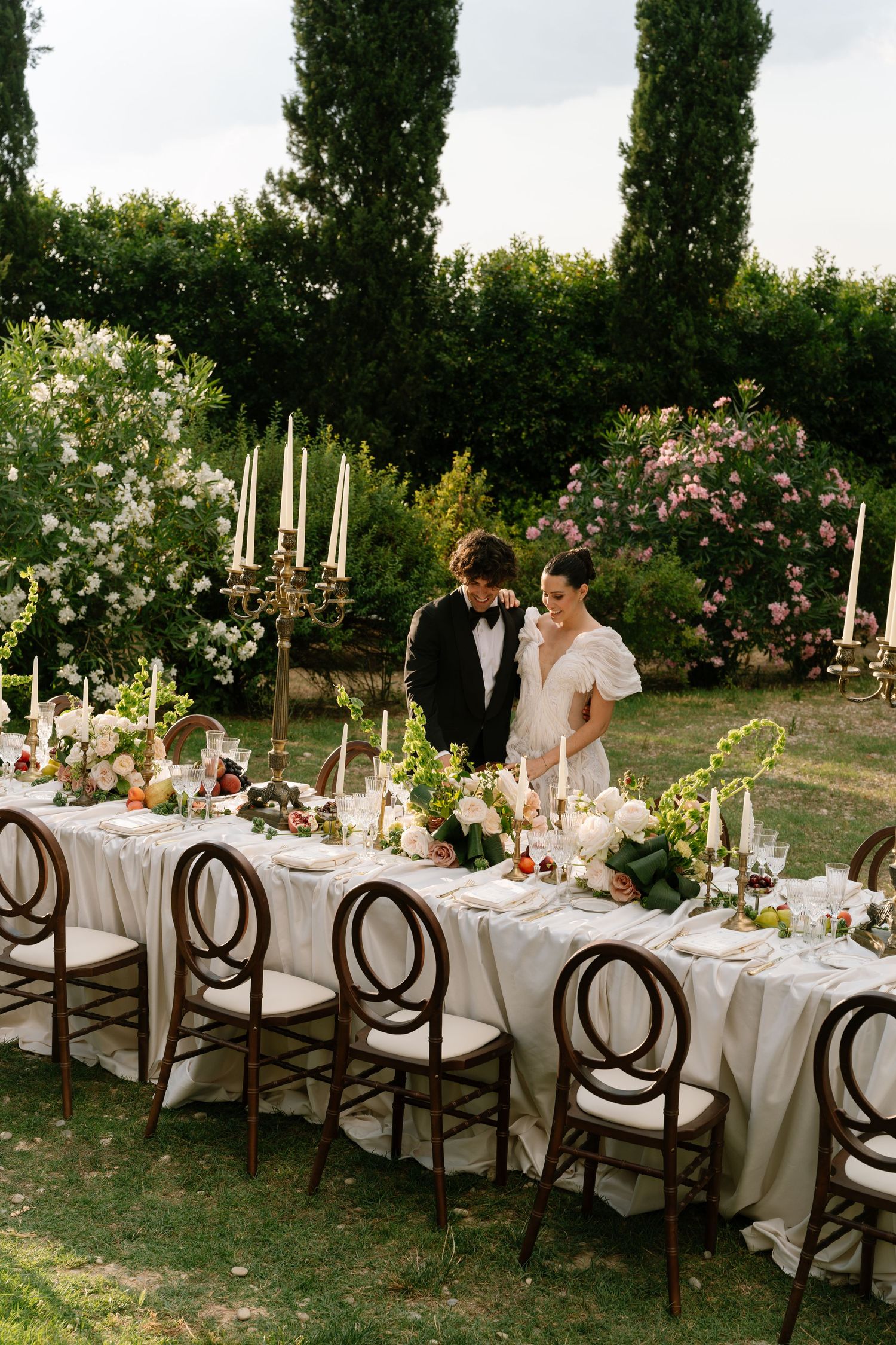 Elegant outdoor wedding reception table with white linens and wooden chairs in a garden setting.
