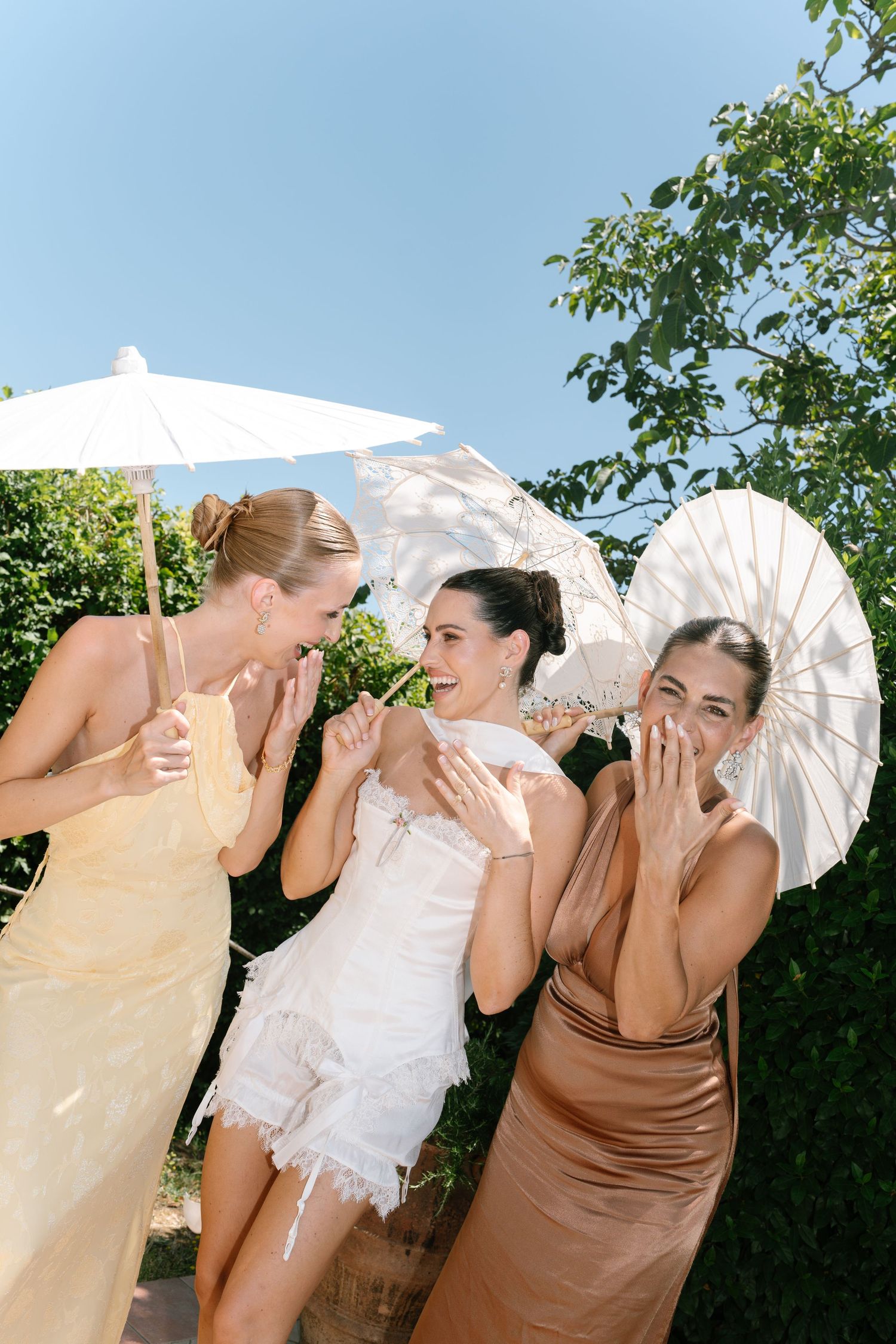 Three women in elegant dresses laugh together while holding white parasols at an outdoor summer event.