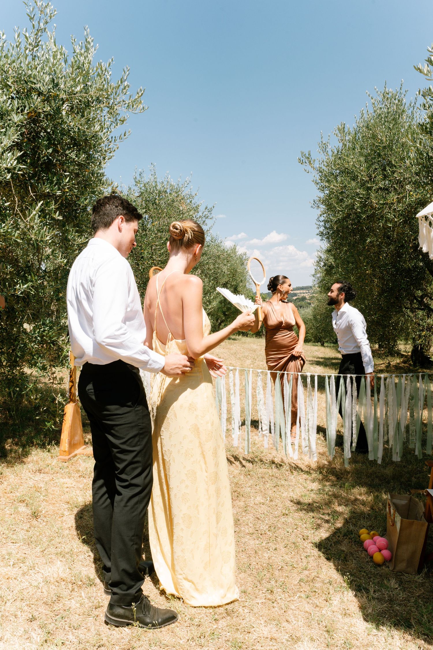 A couple stands among olive trees at an outdoor wedding ceremony with guests in the background.