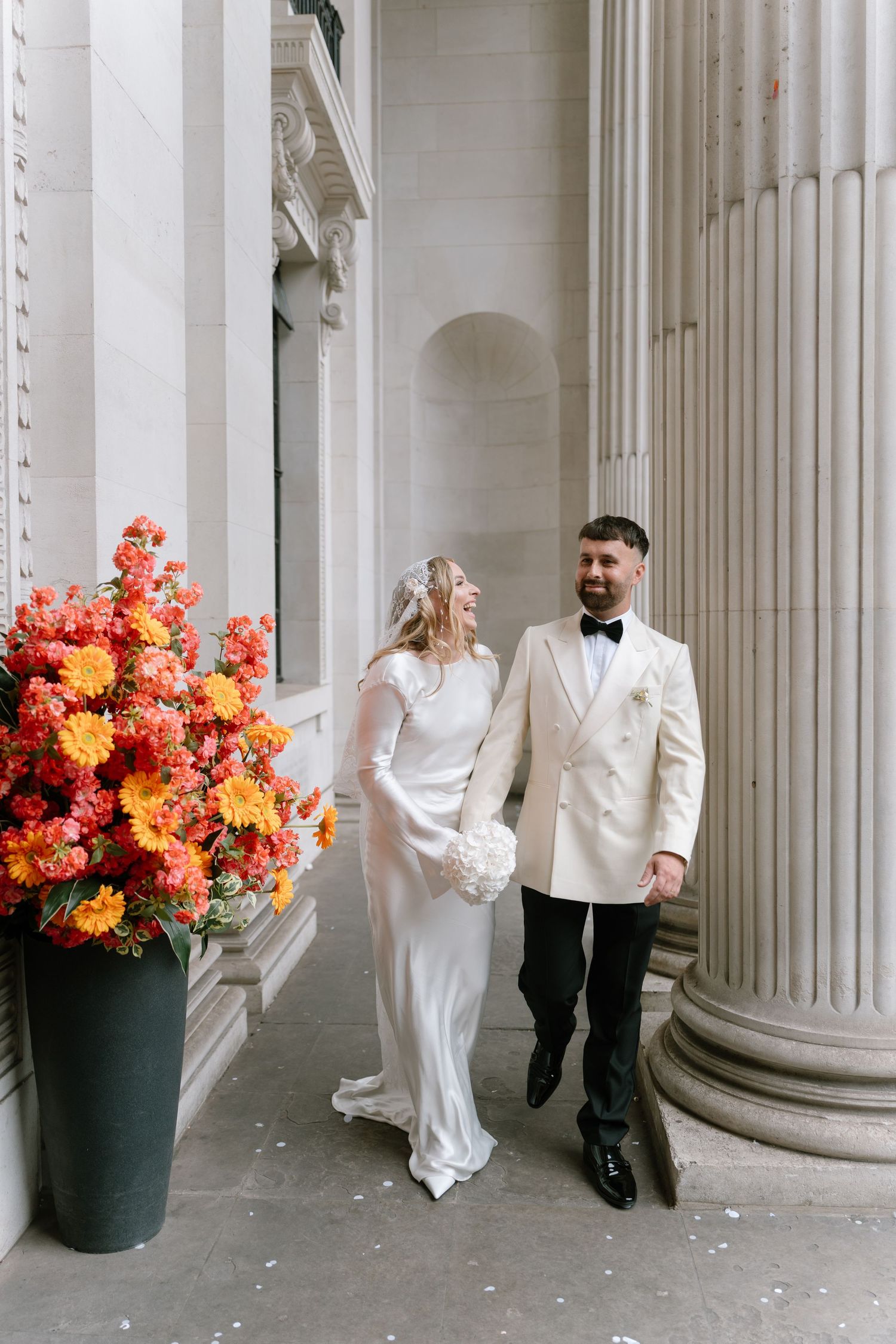 A couple in wedding attire poses playfully between white classical columns with vibrant orange floral arrangements.