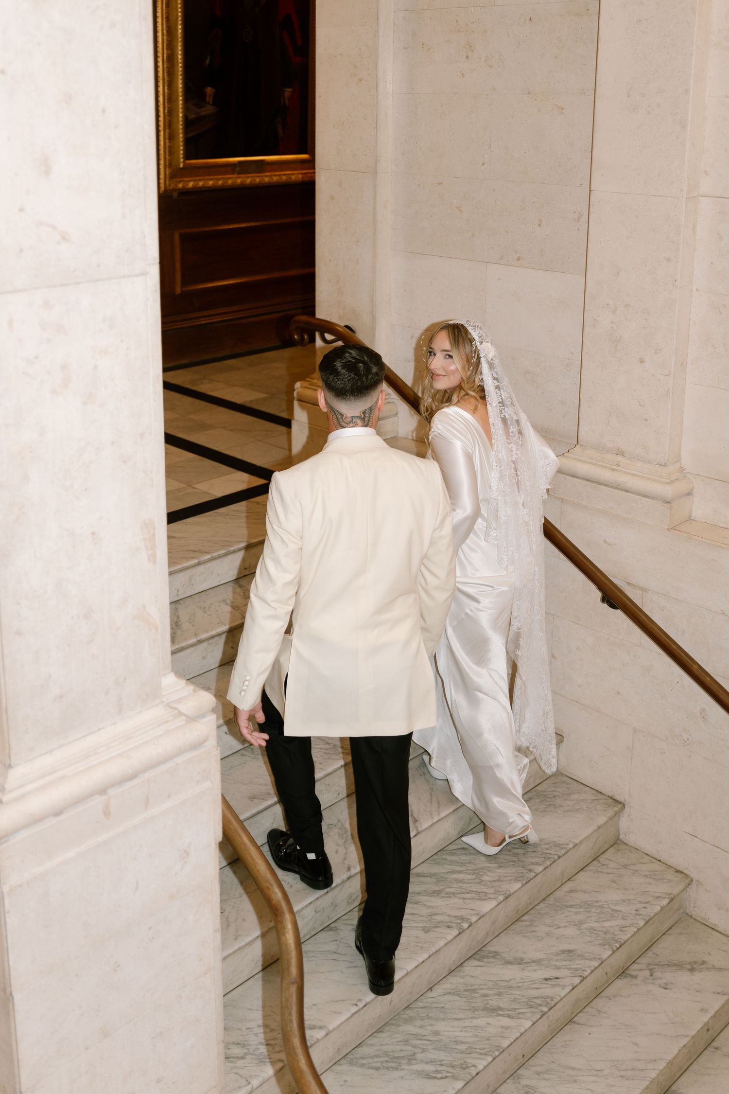 A couple ascends marble stairs in formal white wedding attire.