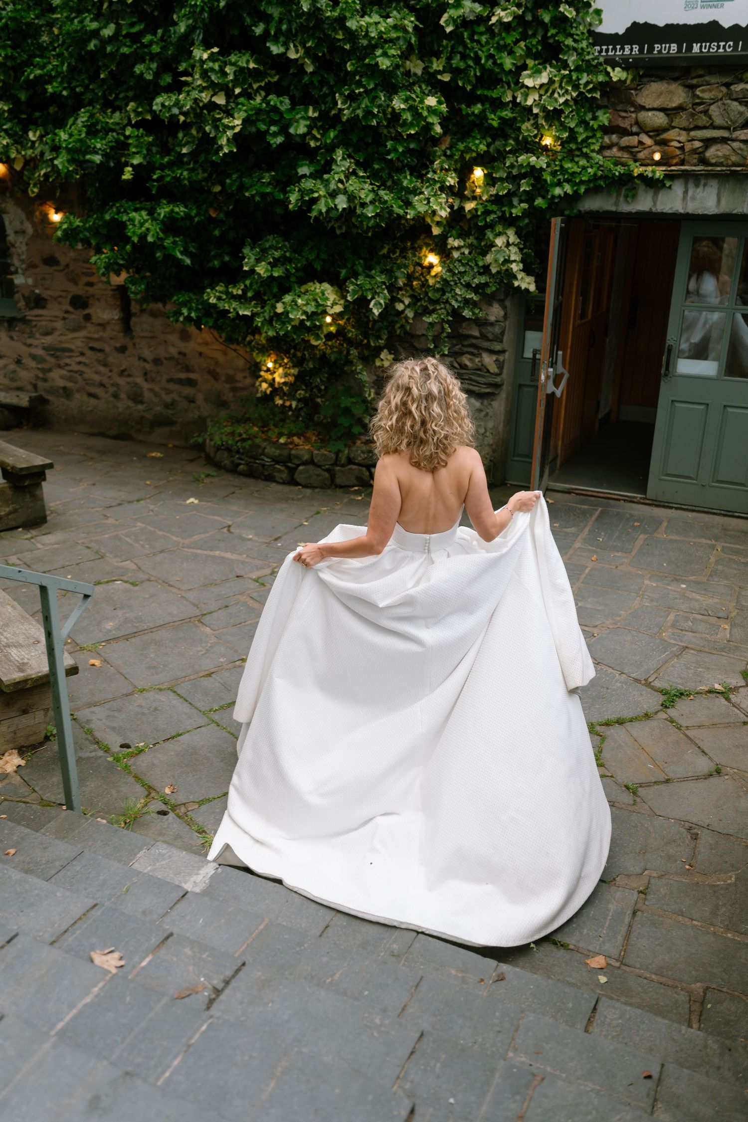 Bride shows off wedding dress train on stone patio surrounded by greenery.